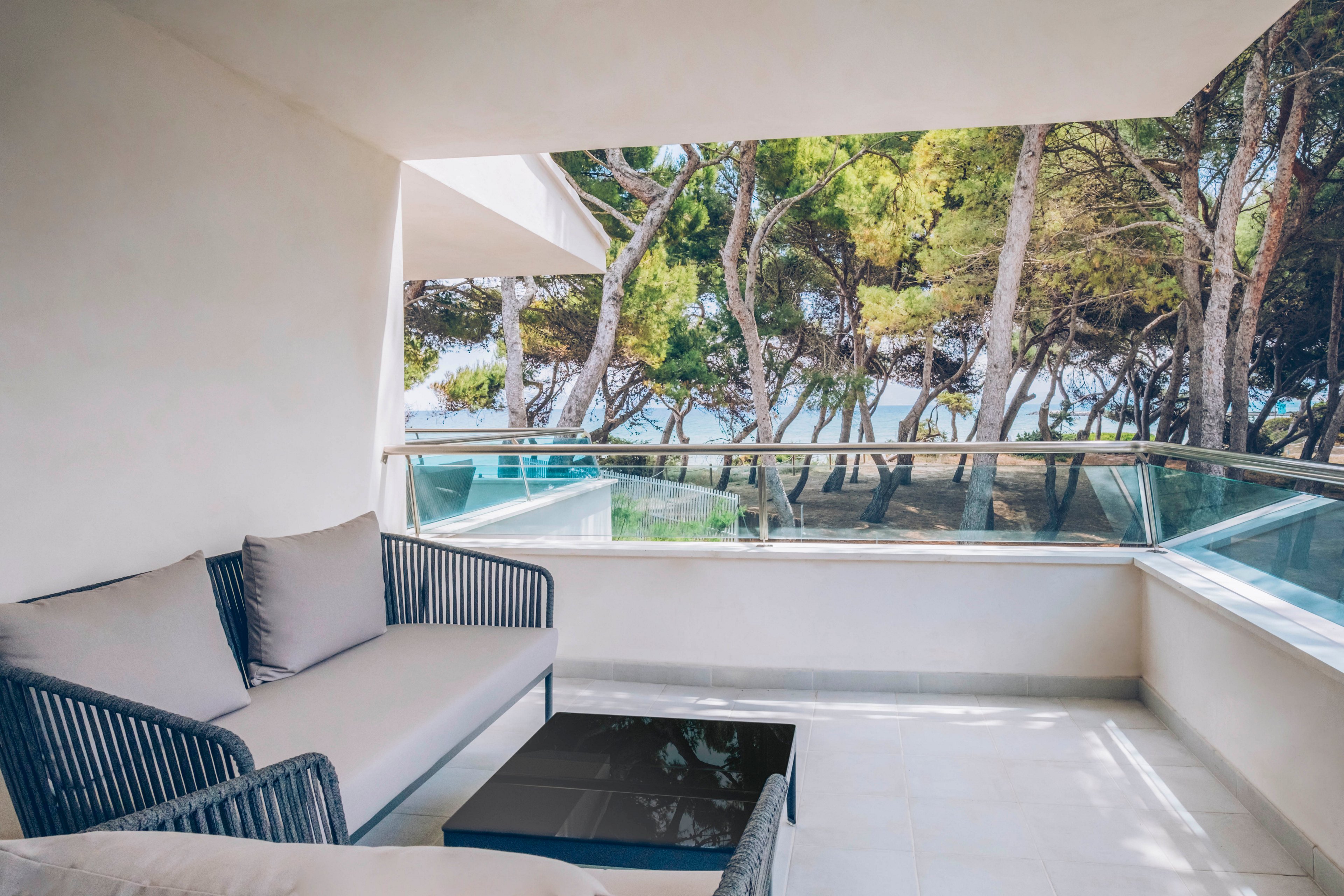 A contemporary balcony lounge features a sleek sofa with neutral cushions and a low black coffee table. Glass railings open the space to a view of tall pine trees and glimpses of the sea beyond. Soft natural light and clean white surfaces create a calm, airy atmosphere. The scene suggests a tranquil vacation retreat or modern seaside apartment.