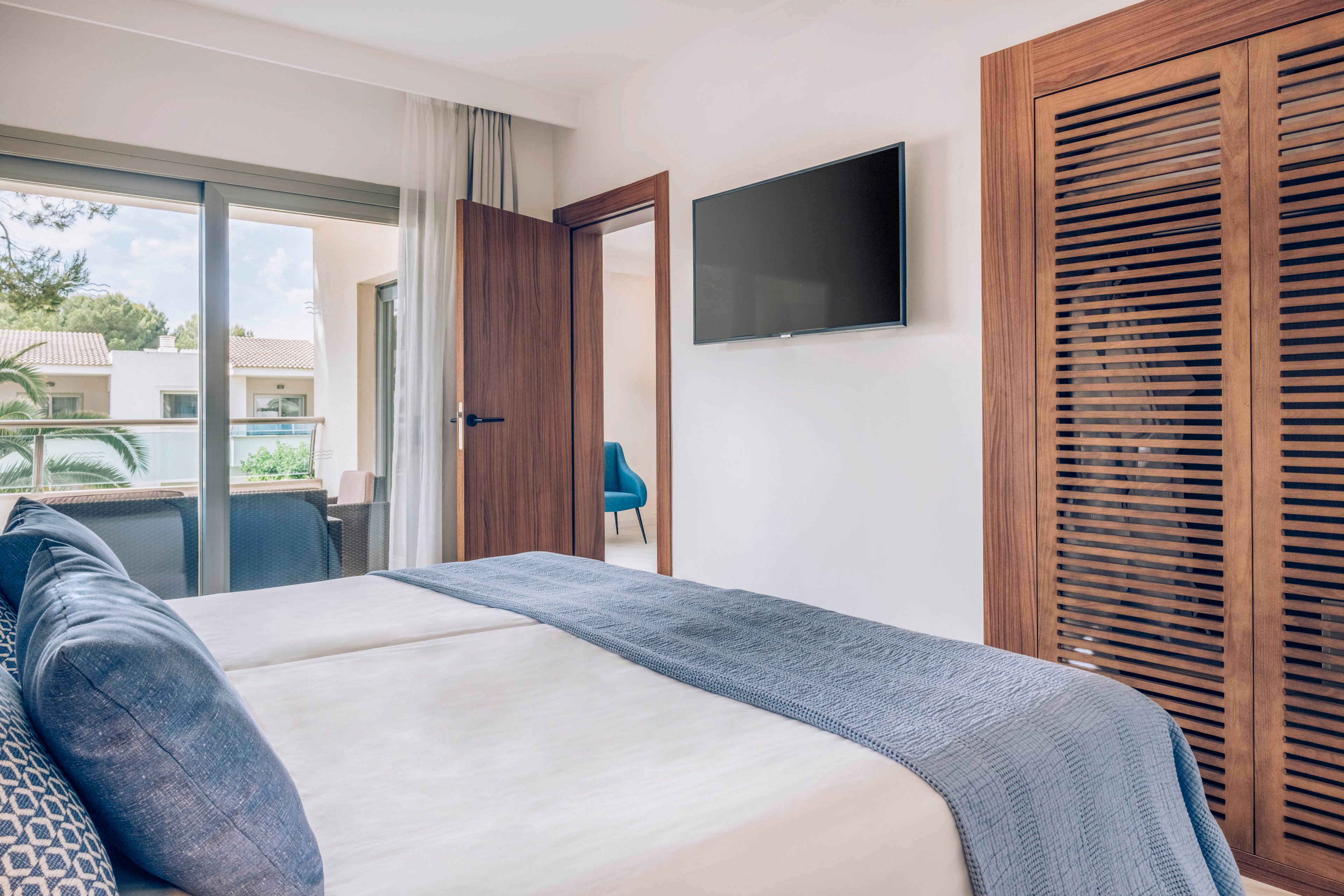 A contemporary bedroom features two neatly made twin beds with blue and white bedding. A flat-screen television is mounted on the wall opposite the beds, and a wooden louvered wardrobe stands nearby. Large sliding glass doors open to a balcony with views of neighboring buildings and greenery. Soft natural light and neutral tones create a calm, inviting atmosphere.