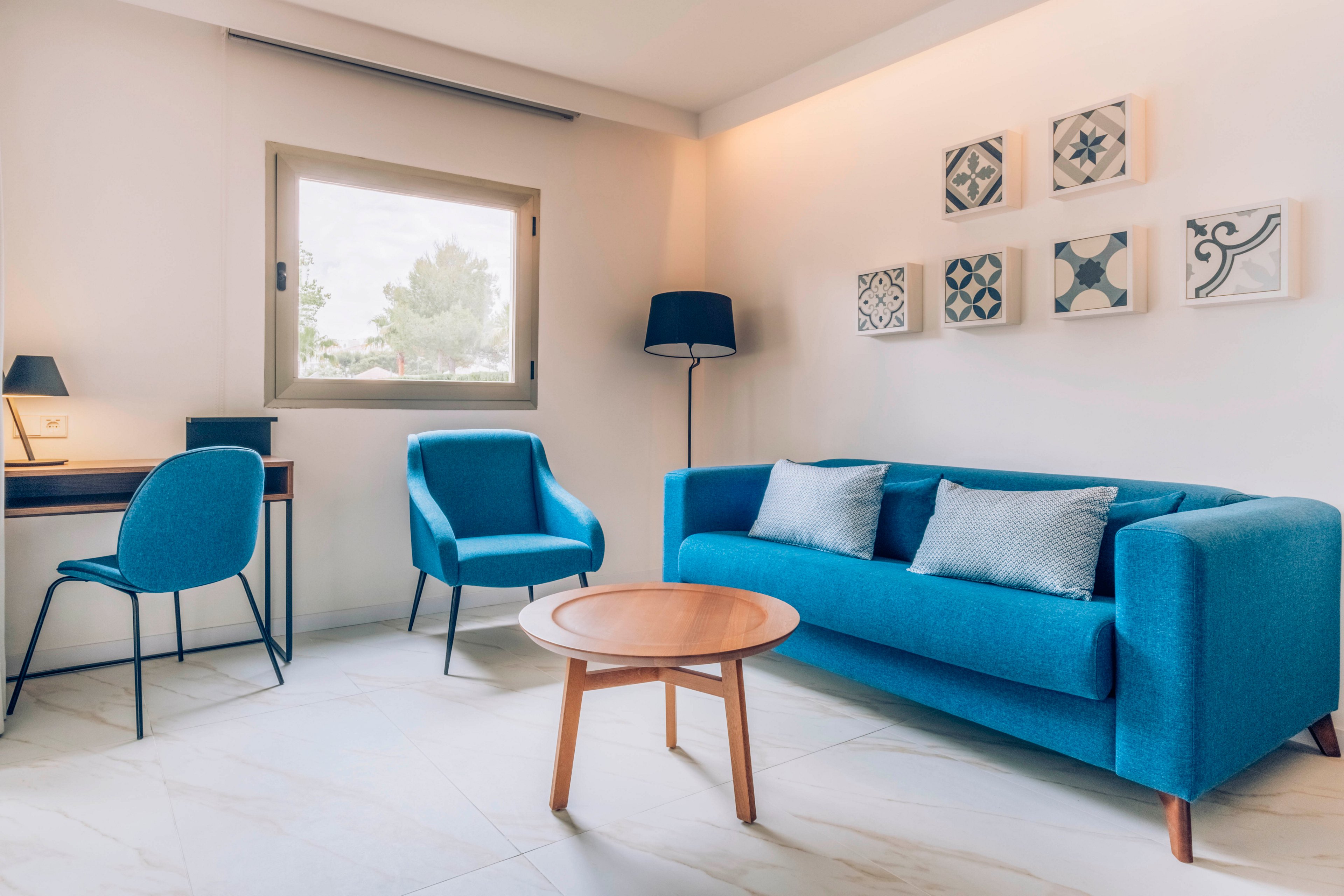 A contemporary living room features a bright blue sofa and matching armchair as the main focal points. The space is styled with a round wooden coffee table, a black floor lamp, and a compact desk with a chair near the window. Soft natural light enters through the window, illuminating the light-colored walls and floor. Decorative patterned tiles hang on the wall, adding subtle artistic detail to the minimalist interior.