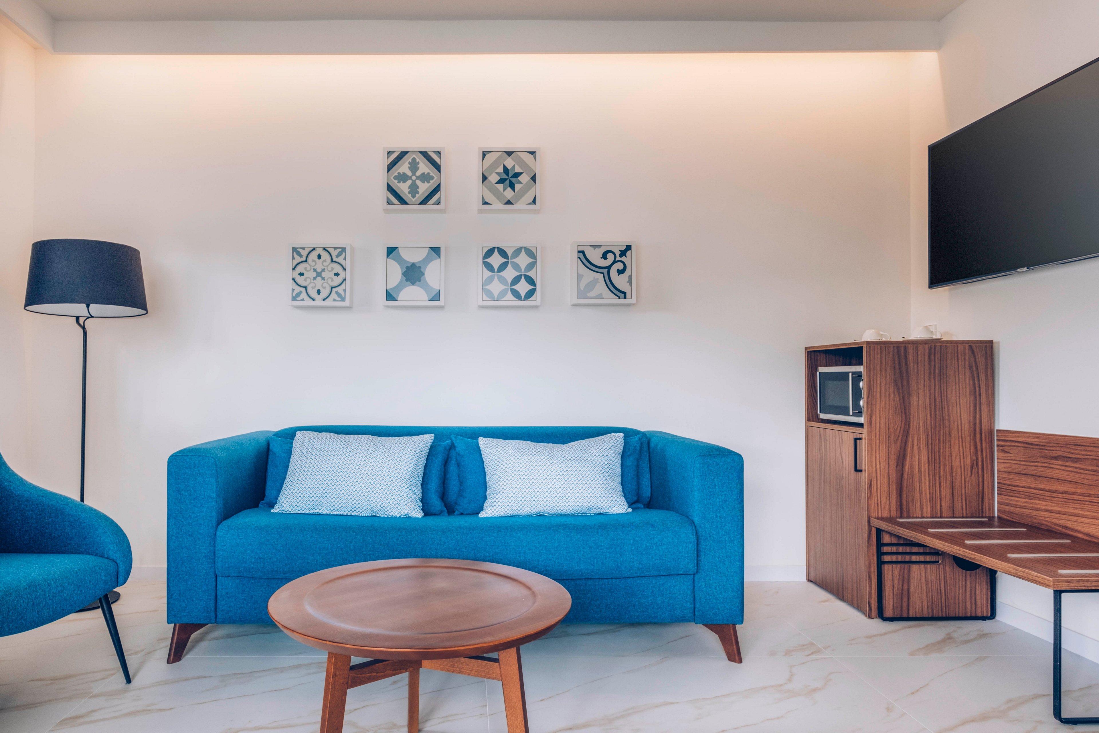 A contemporary living room features a bright blue sofa with matching cushions as the central focus. The space includes a round wooden coffee table, a blue accent chair, and a tall floor lamp on one side. On the wall above the sofa, six decorative tiles with geometric patterns add visual interest. To the right, a wooden cabinet with a microwave and a mounted flat-screen television complete the clean, minimalist interior.