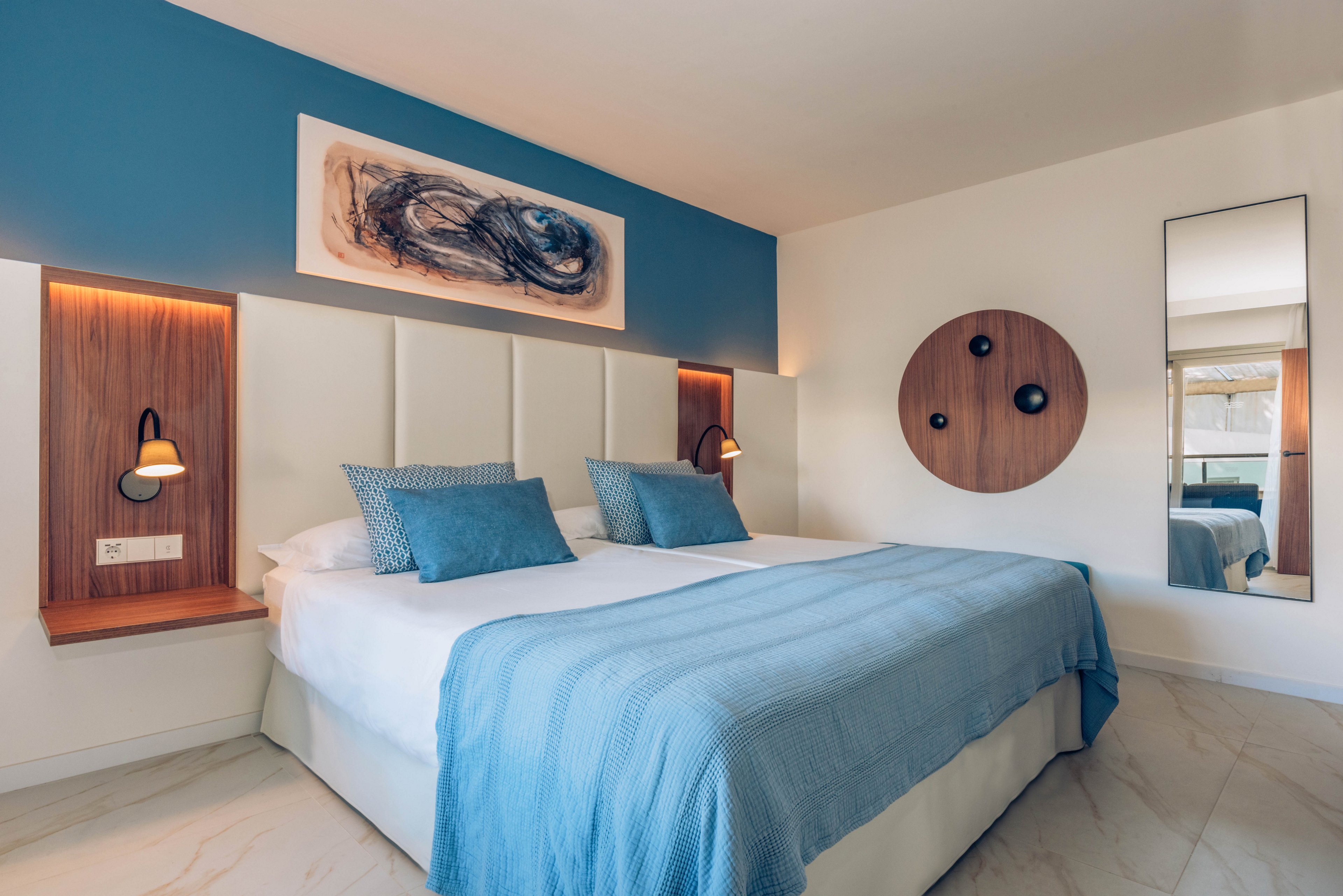 A contemporary double bedroom features a large bed with white linens and light blue bedding. The room has a blue accent wall with integrated wooden headboard panels and built-in reading lights. Minimalist artwork and circular wooden wall decor add visual interest, while a tall mirror reflects the bright, airy space. The overall design feels clean, modern, and inviting.