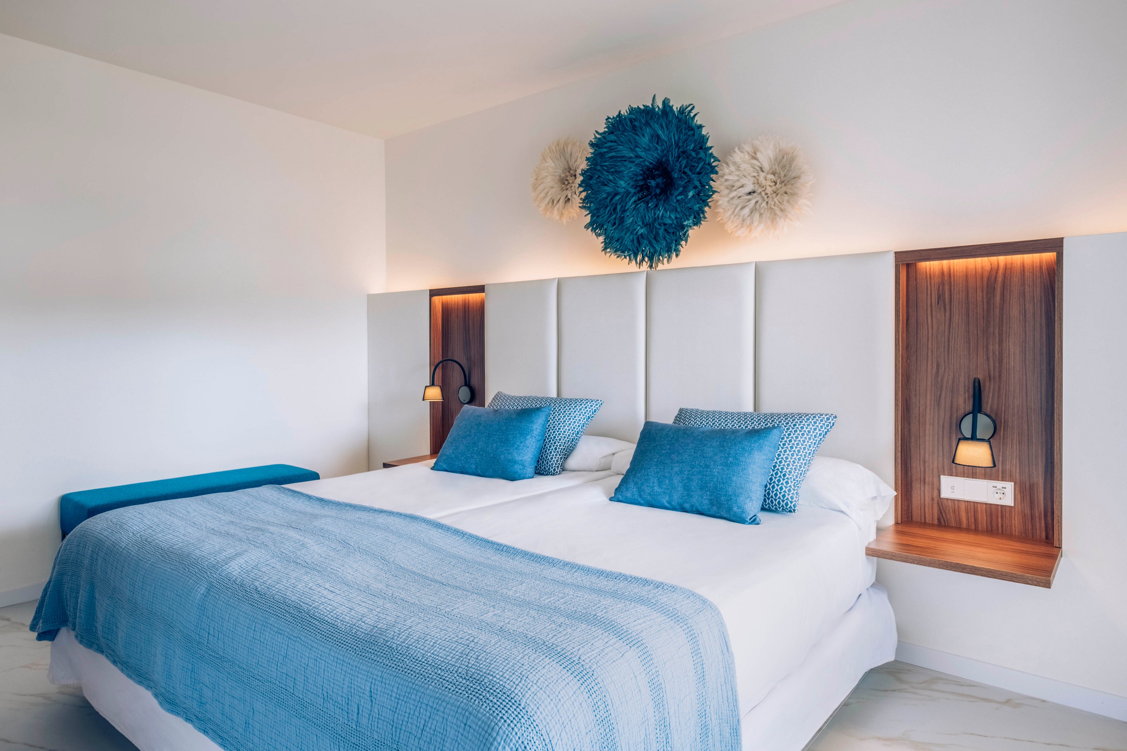 A neatly made double bed with a blue duvet and matching pillows is the focal point of this modern bedroom. The room features a minimalist design with white walls, padded headboard panels, and warm wooden bedside niches. Soft lighting from built-in bedside lamps creates a cozy atmosphere. Decorative wall art above the headboard and a blue bench at the foot of the bed add visual interest and color contrast.