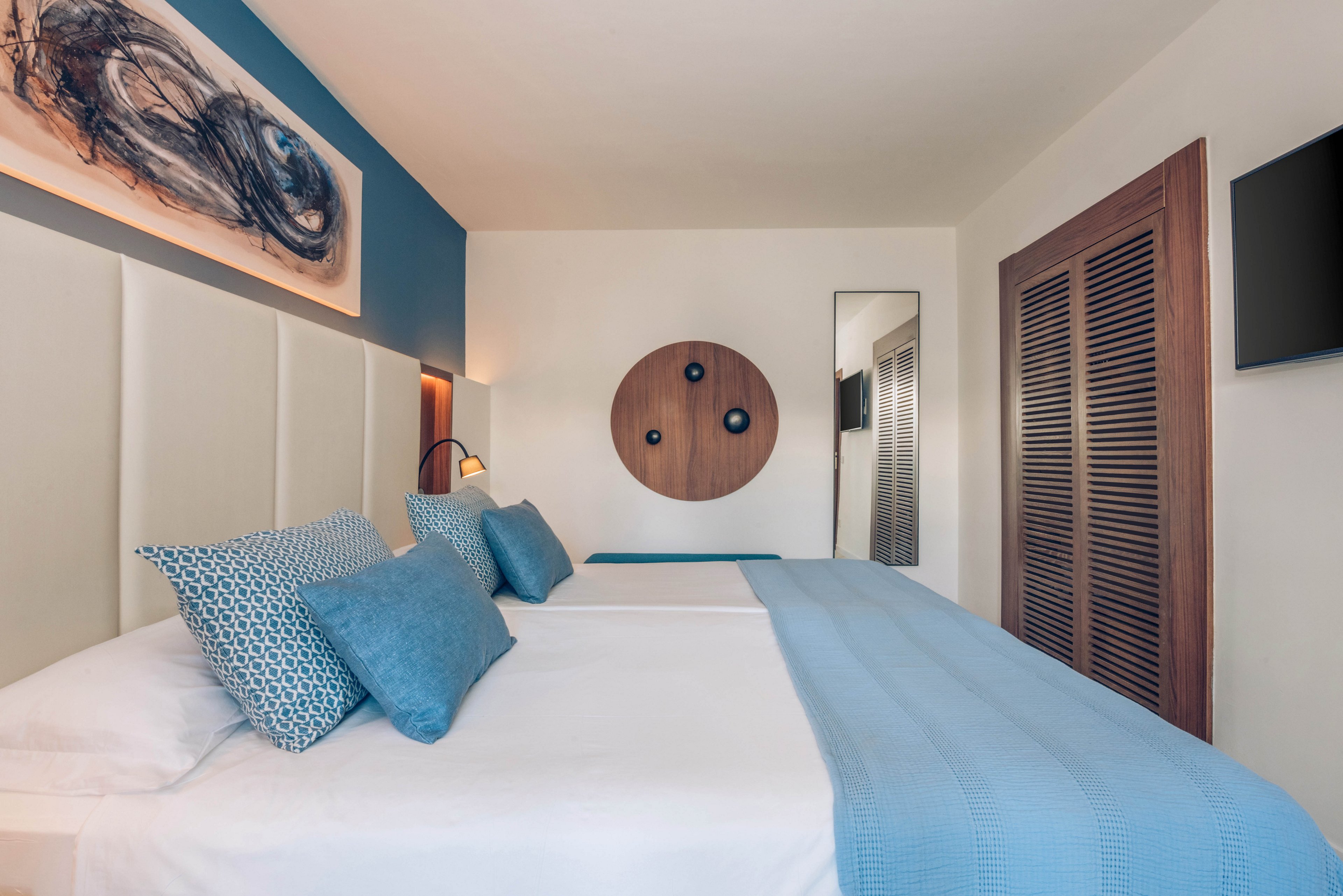 A contemporary hotel bedroom features a large neatly made bed with white linens and blue pillows and throw. The room has a minimalist design with a padded headboard, wooden closet doors, and a wall-mounted television. Decorative artwork, including an abstract painting and a round wooden wall piece, add visual interest. Soft lighting and a neutral color palette create a calm, inviting atmosphere.