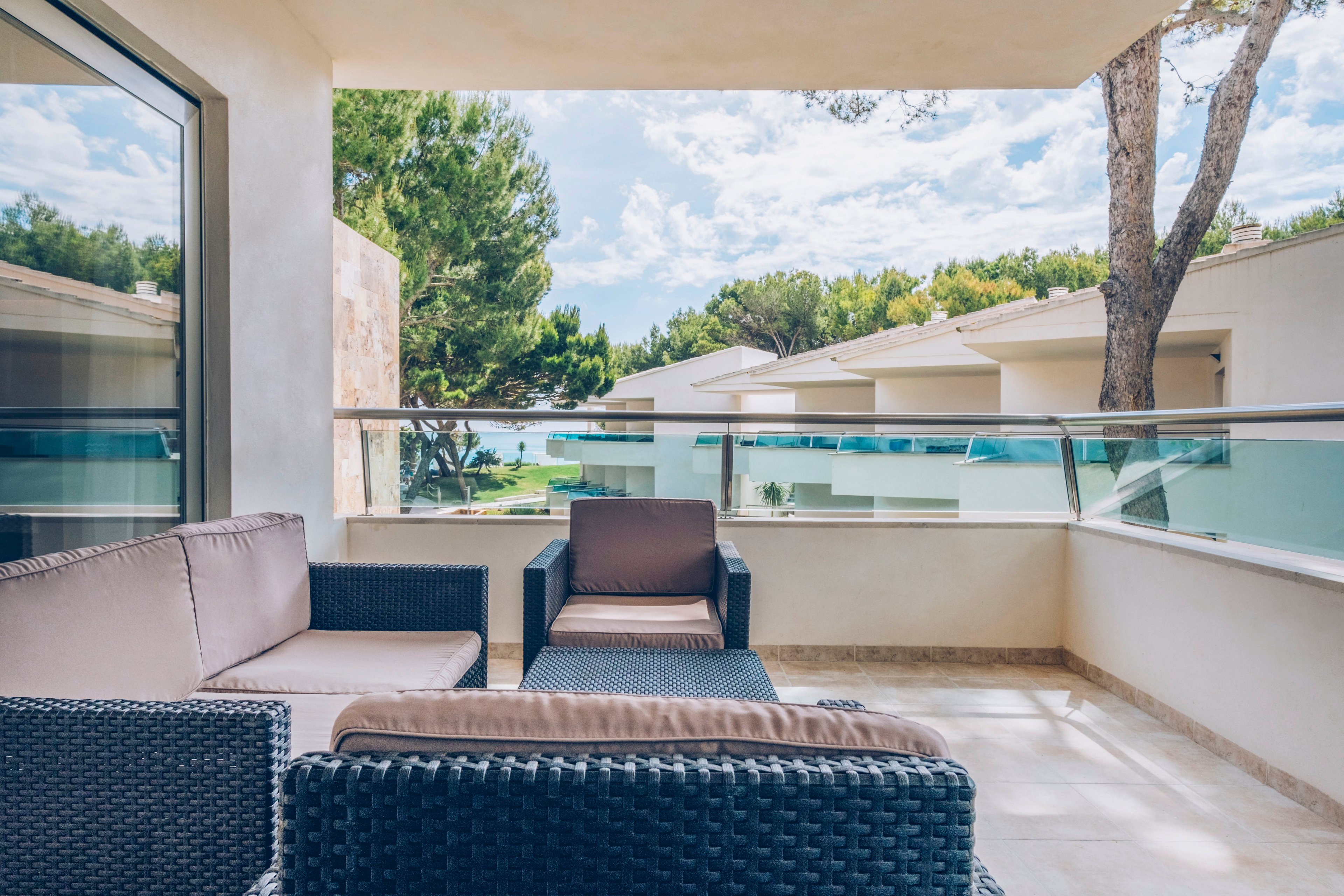 A modern balcony features woven rattan-style furniture with cushioned seating. The terrace overlooks a contemporary resort complex with white buildings and flat roofs. Glass railings and light stone flooring create an open, airy feel. Trees and a glimpse of water in the distance add a relaxing, vacation atmosphere.