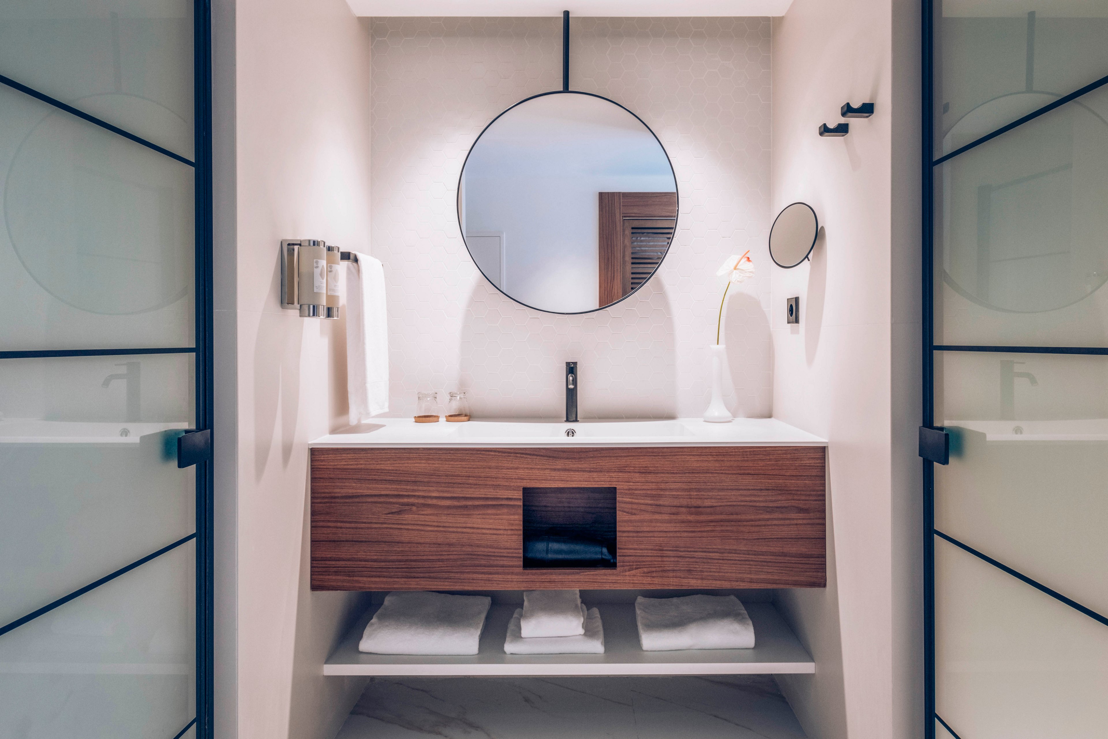 A contemporary bathroom vanity features a sleek wooden cabinet with a white countertop and integrated sink. A round mirror hangs above the faucet, reflecting a doorway behind the viewer. Towels are neatly folded on the open shelf below, and a wall-mounted dispenser and towel ring sit to the left of the sink. Soft, neutral lighting and clean lines create a calm, minimalist atmosphere.