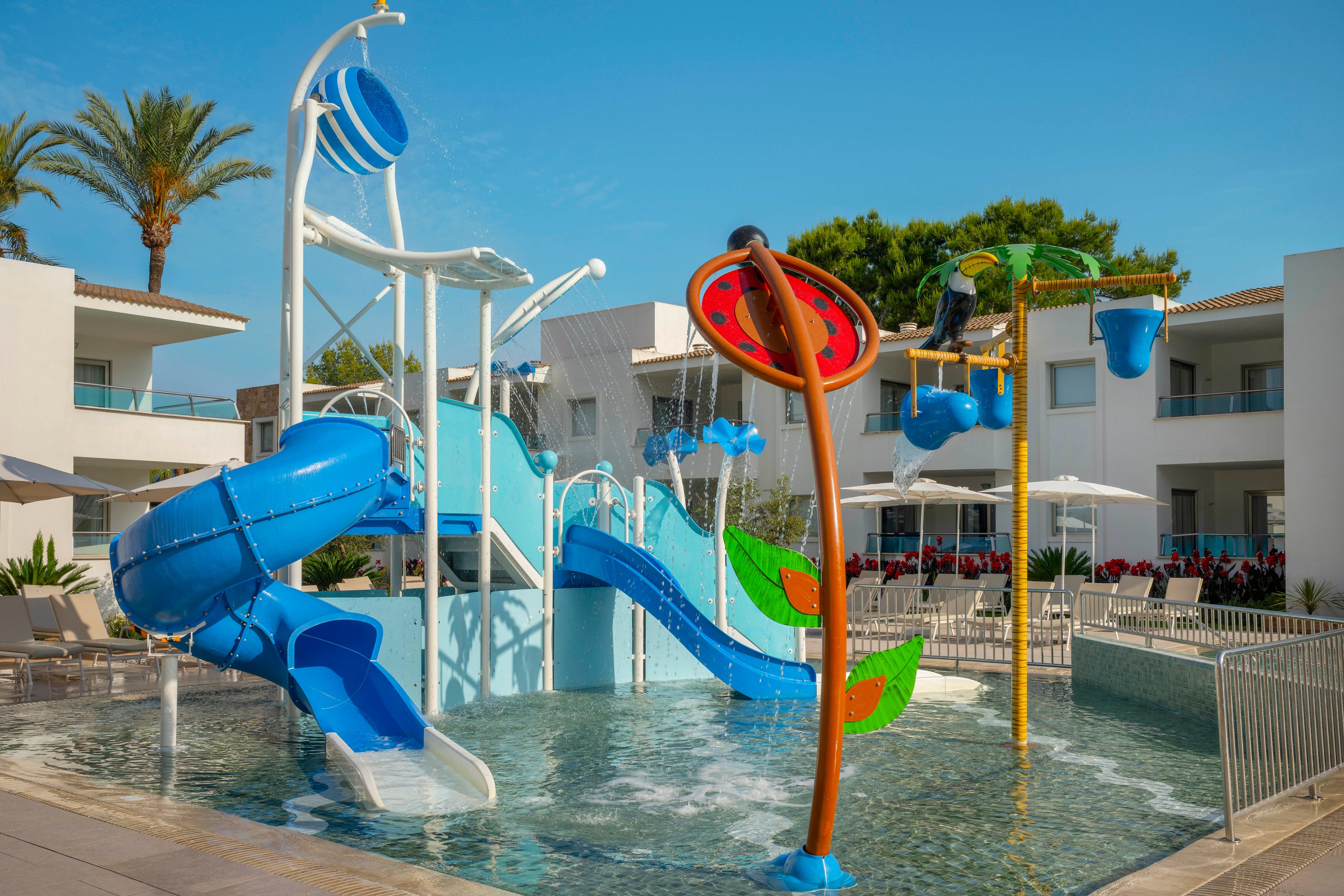 A vibrant outdoor water playground features multiple blue slides and interactive splash structures in a shallow pool. The play area includes tipping buckets, water sprayers, and climbing elements designed for children. Modern white resort buildings and palm trees surround the pool under a clear blue sky, suggesting a warm vacation setting. The scene feels playful, family-friendly, and ideal for leisure at a holiday resort.