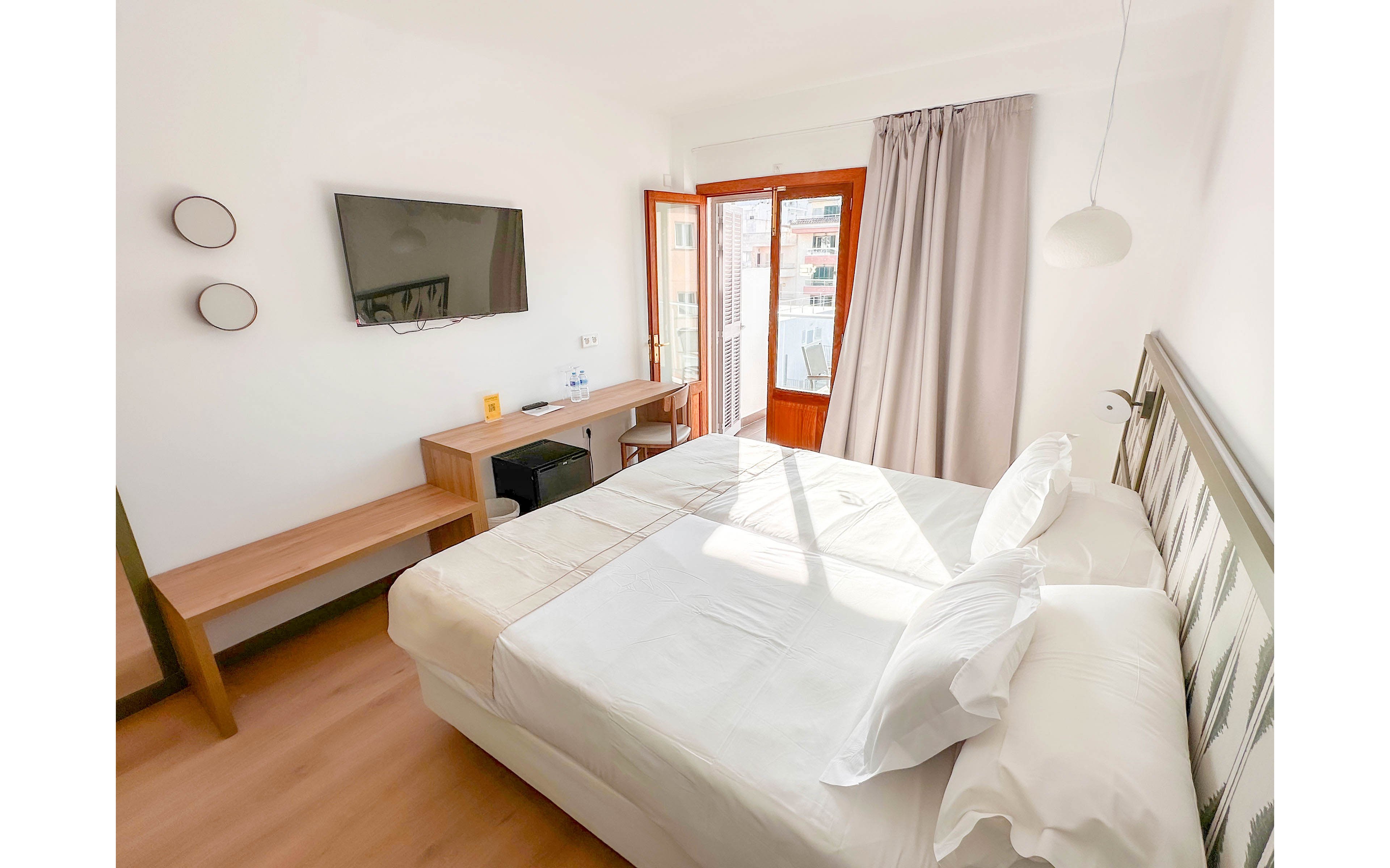 A sunlit hotel bedroom features a neatly made double bed with white linens and a light wood floor. A wall-mounted flat-screen TV faces a simple wooden desk and bench, with a chair tucked underneath. Large glass doors open to a balcony, letting in natural light and views of nearby buildings. Neutral curtains and minimal decor create a clean, contemporary atmosphere.