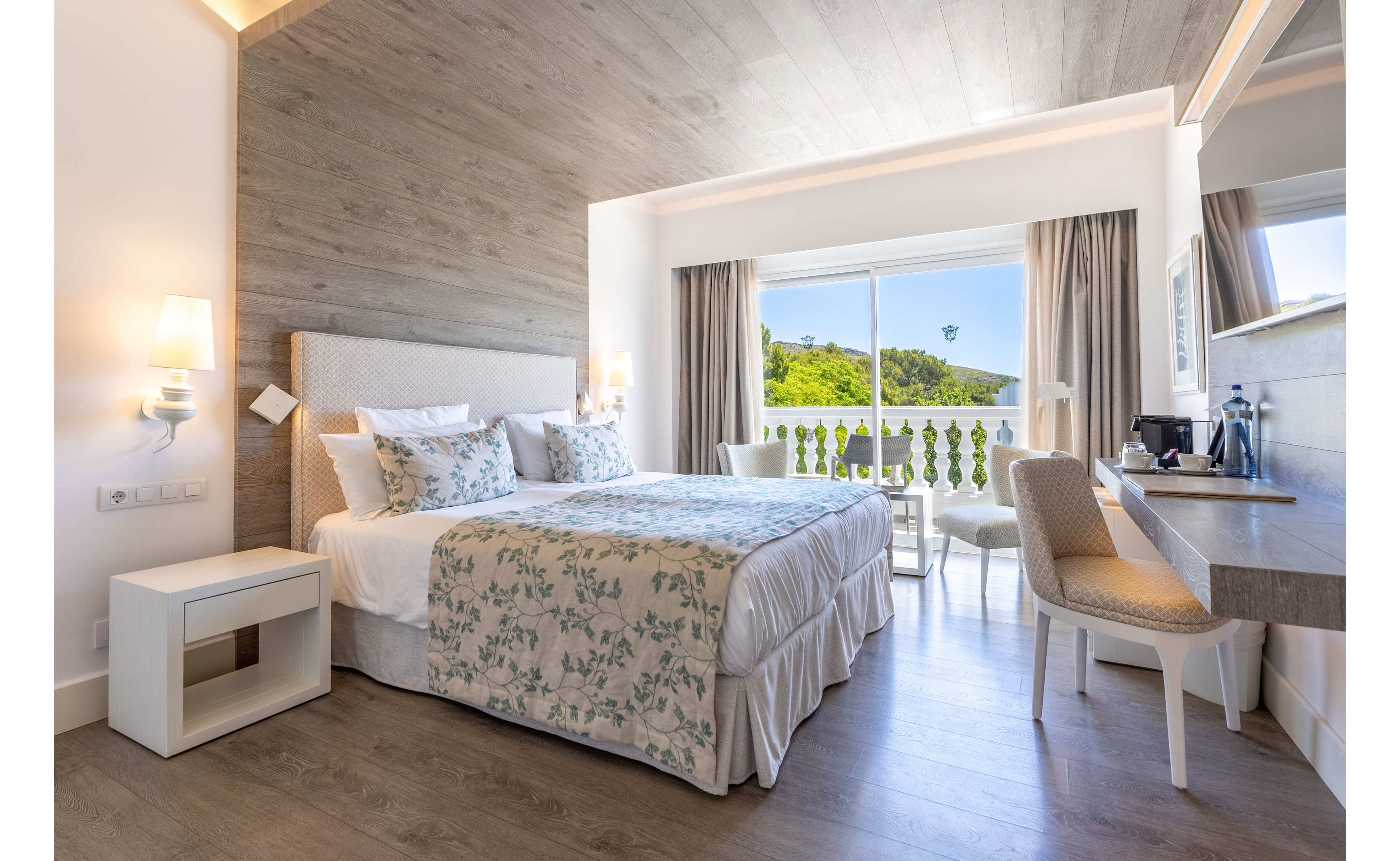 A modern hotel bedroom features a large double bed with floral bedding and multiple pillows. Light wood floors and a wood-paneled ceiling create a warm, contemporary atmosphere. A desk with a chair, coffee station, and wall-mounted lamps add functional comfort. Large sliding glass doors open to a balcony with white railings and a scenic view of green hills under a clear sky.