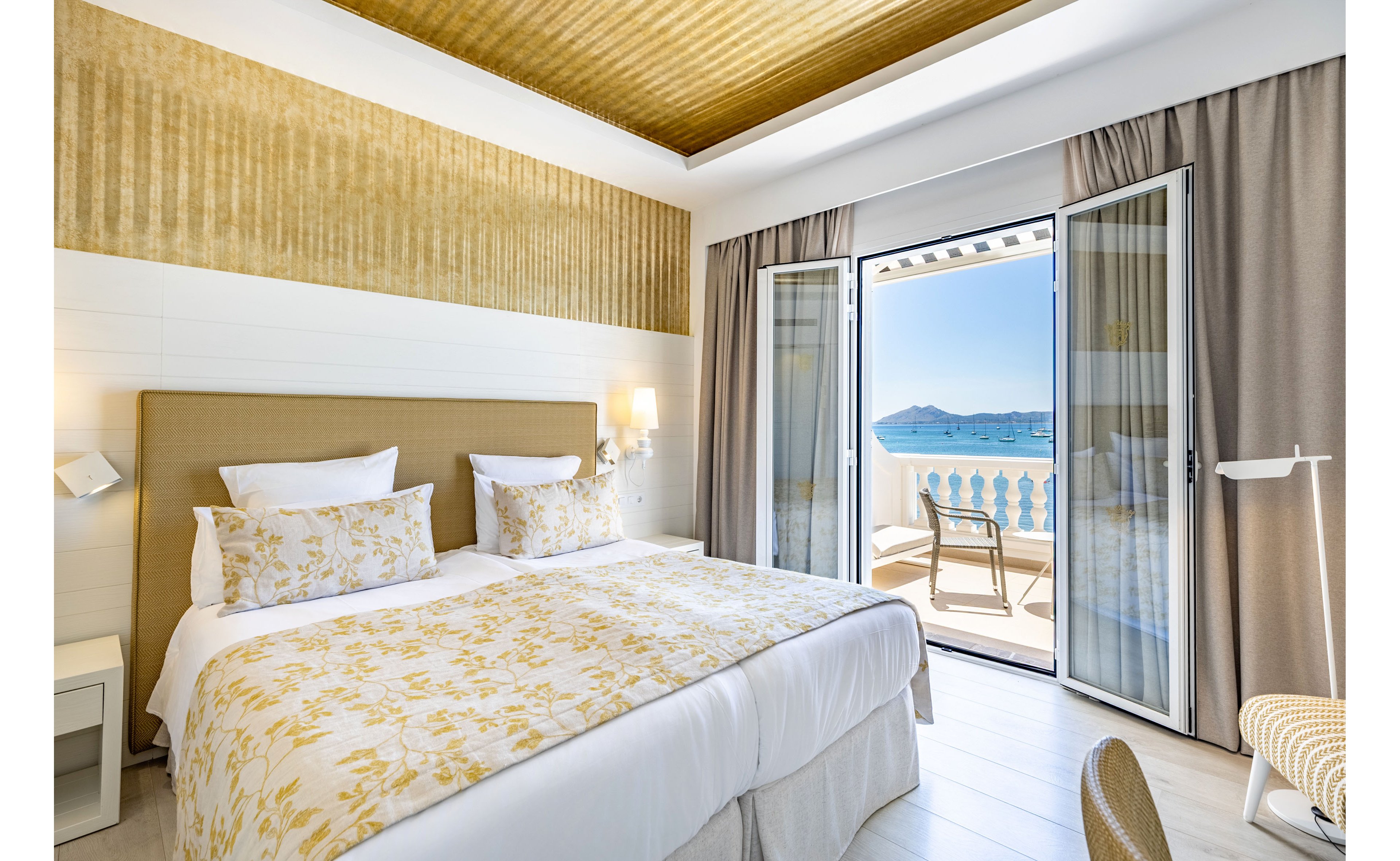 A modern hotel bedroom features a large neatly made bed with patterned yellow and white bedding. Neutral-toned walls, curtains, and a textured golden ceiling create a warm, contemporary atmosphere. Open glass doors lead to a balcony with a seating area and a scenic view of the sea and distant mountains. Natural daylight fills the room, highlighting the clean, inviting interior design.