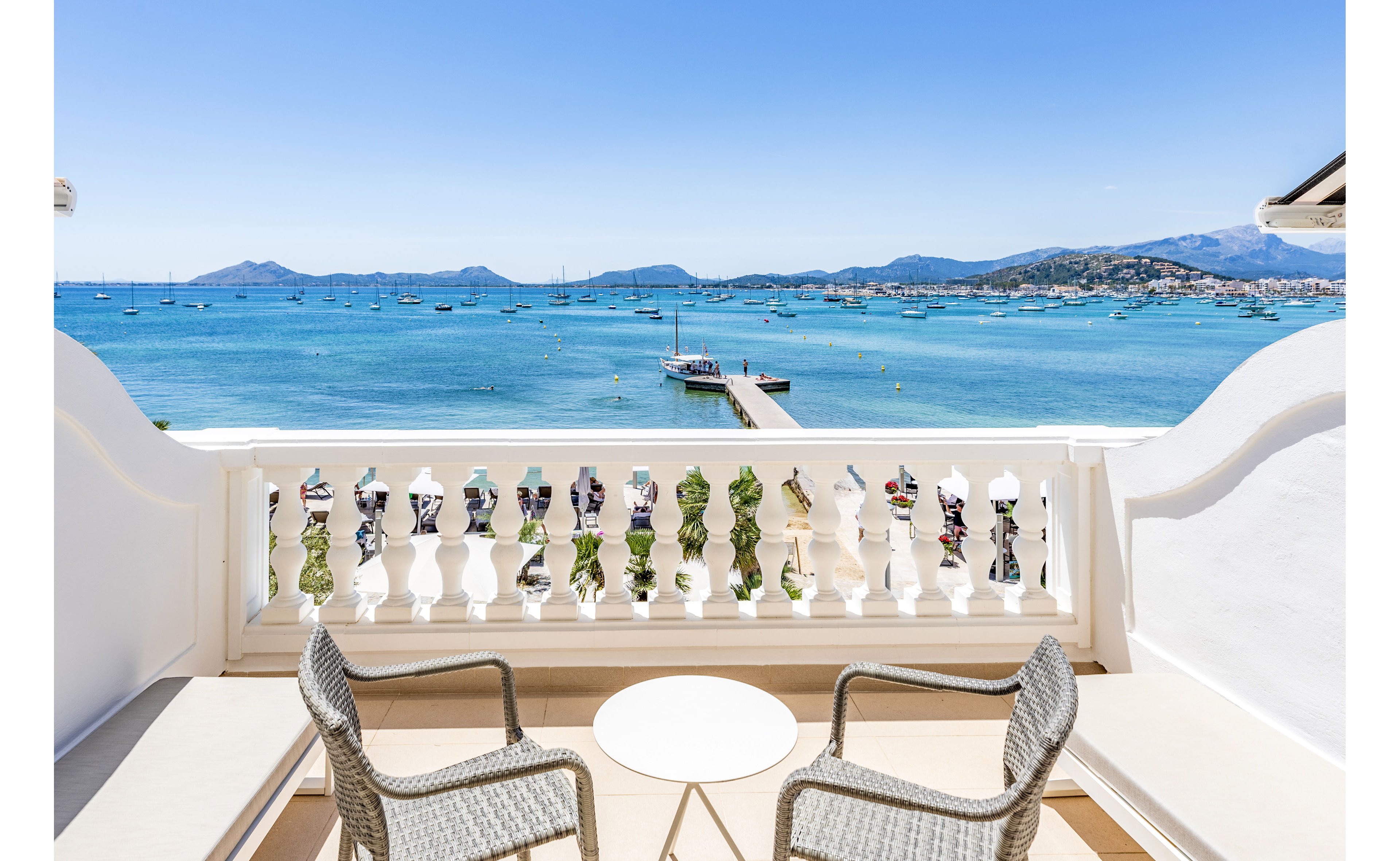 A bright seaside terrace features two wicker chairs and a small round table facing a white balustrade. Beyond the terrace, a wooden pier extends into calm turquoise water dotted with anchored boats. Distant hills and a coastal town line the horizon under a clear blue sky. The scene suggests a peaceful vacation setting with a focus on relaxation and ocean views.
