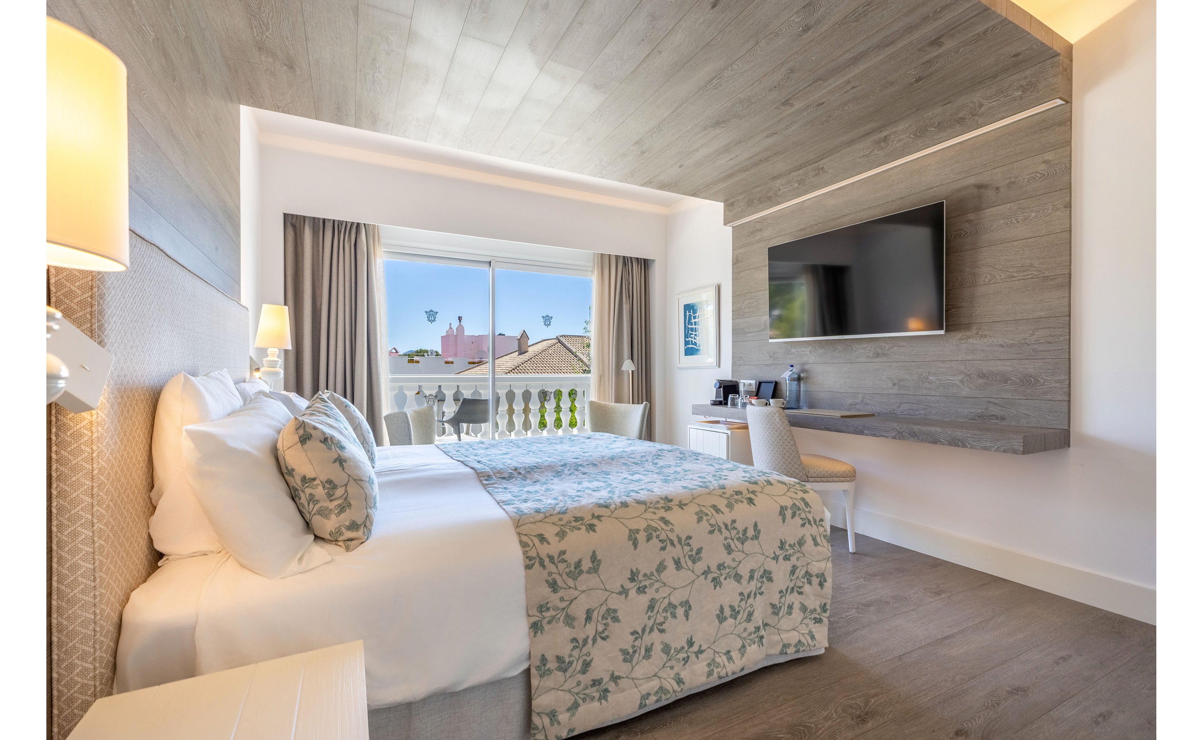 A bright, modern hotel bedroom features a neatly made double bed with floral bedding and multiple pillows. The room includes a large wall-mounted flat-screen TV opposite the bed and a small desk with a chair and coffee station. Light wood flooring and a wood-paneled ceiling create a warm, contemporary atmosphere. A sliding glass door opens to a balcony with white balustrades and a view of rooftops and cable cars against a clear blue sky.