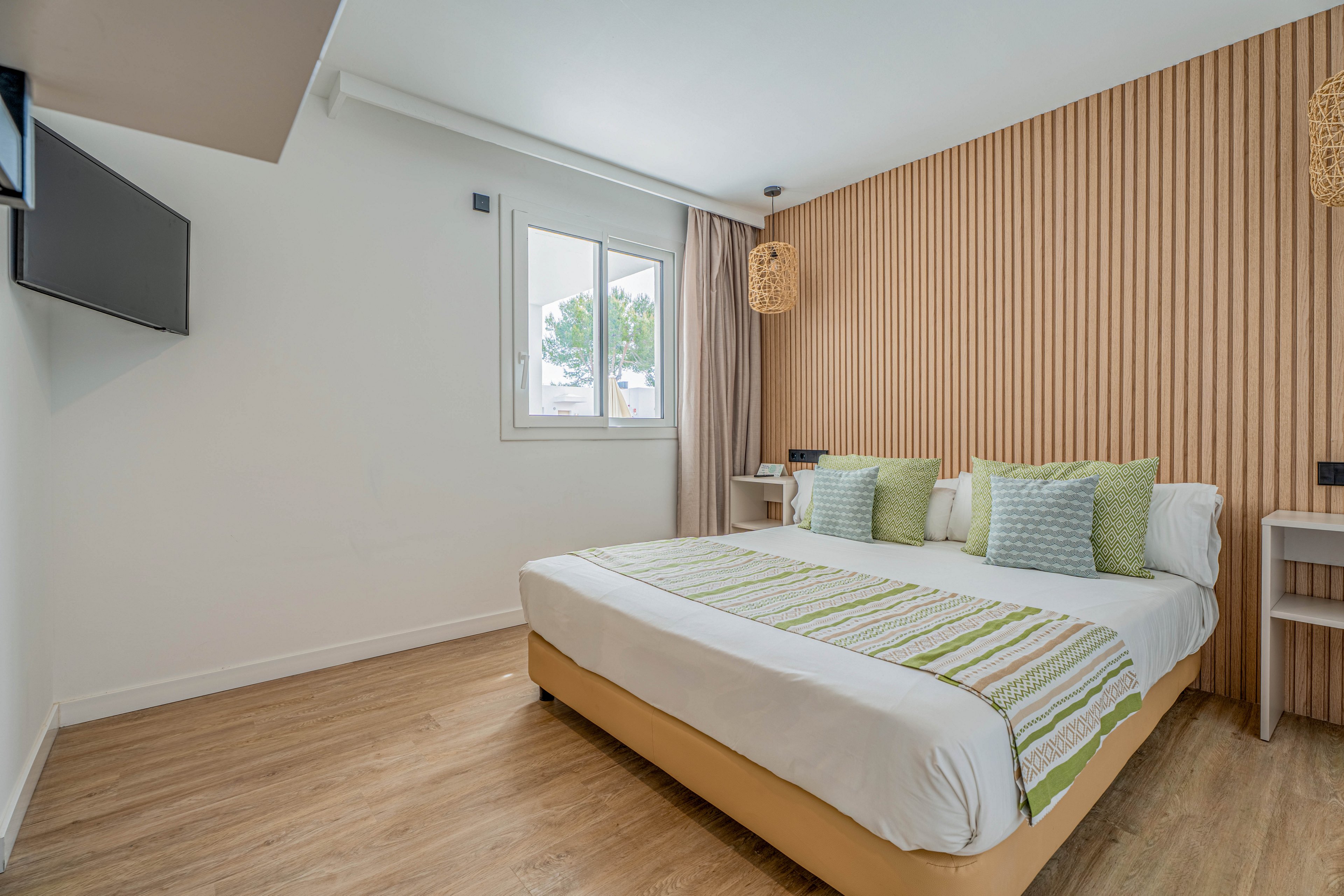 Modern bedroom interior featuring a neatly made double bed with striped green and white textiles and multiple decorative pillows. The room has light wooden flooring, a slatted wood accent wall, and two hanging woven pendant lamps. A wall-mounted flat-screen TV faces the bed, and a window with beige curtains lets in natural daylight. The overall design is clean, minimalist, and inviting, with soft neutral tones and subtle green accents.