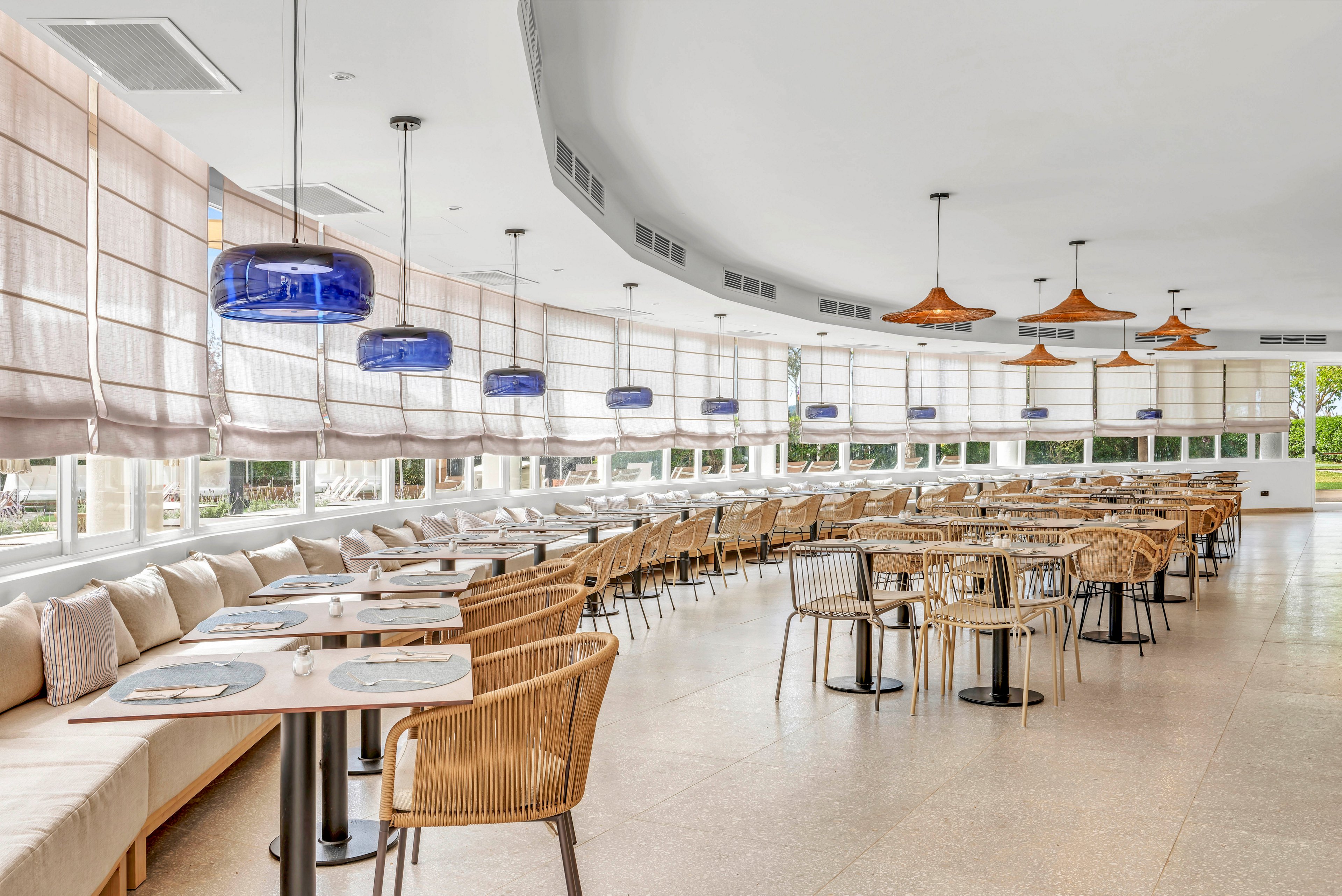 Spacious contemporary restaurant dining area with neatly arranged tables and chairs. Long banquette seating with cushions lines the wall beneath large windows covered by sheer blinds. Blue and copper pendant lights hang from the curved ceiling, adding color accents to the neutral palette. The room is empty of people, emphasizing clean lines, natural light, and an airy atmosphere.