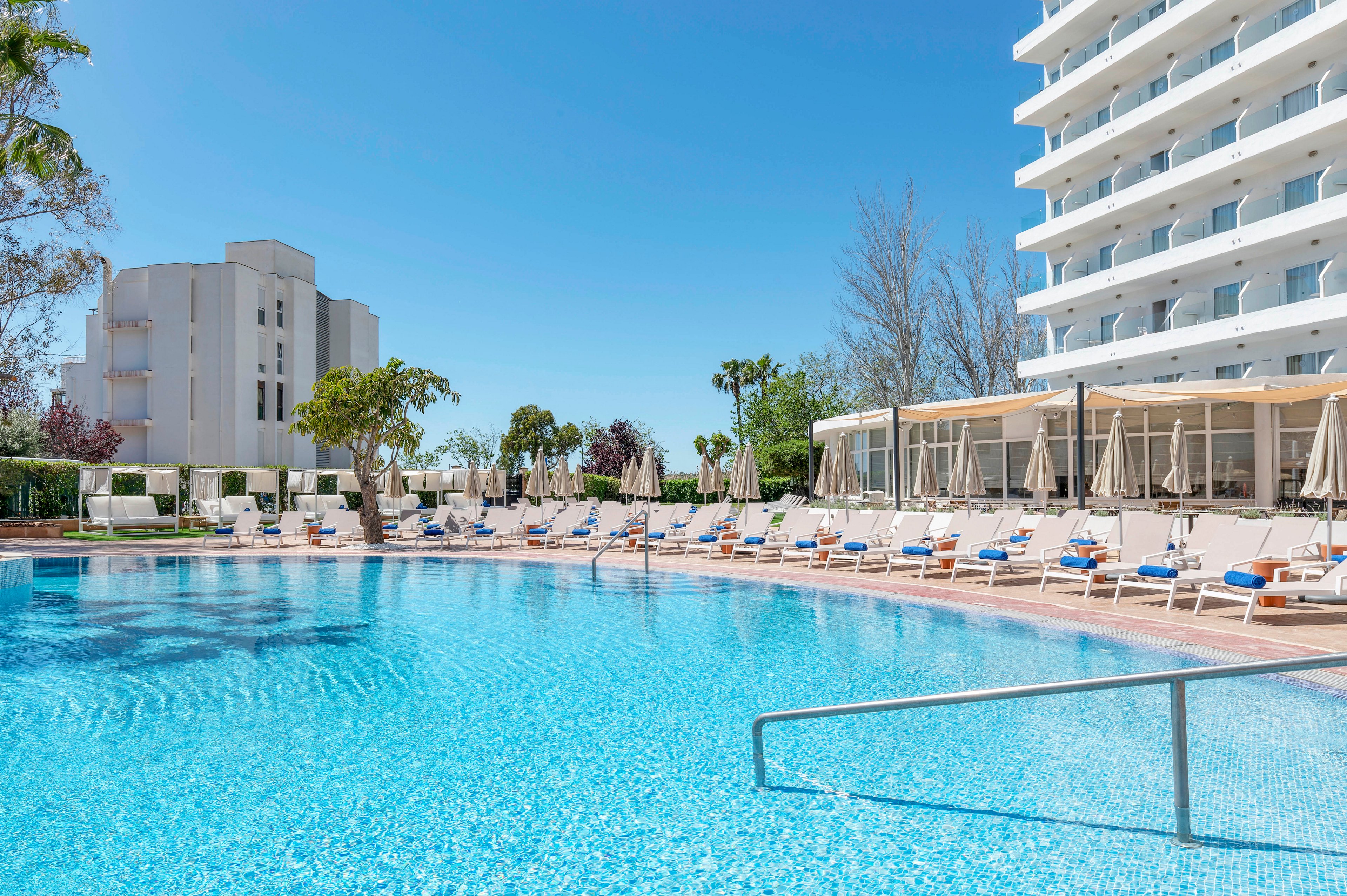 A large outdoor swimming pool sparkles in front of a modern multi-story resort hotel. Sun loungers with umbrellas line the poolside terrace, suggesting a relaxing vacation setting. The sky is clear and blue, and surrounding trees and landscaping add a touch of greenery. The overall scene conveys a bright, sunny day at a contemporary holiday destination.