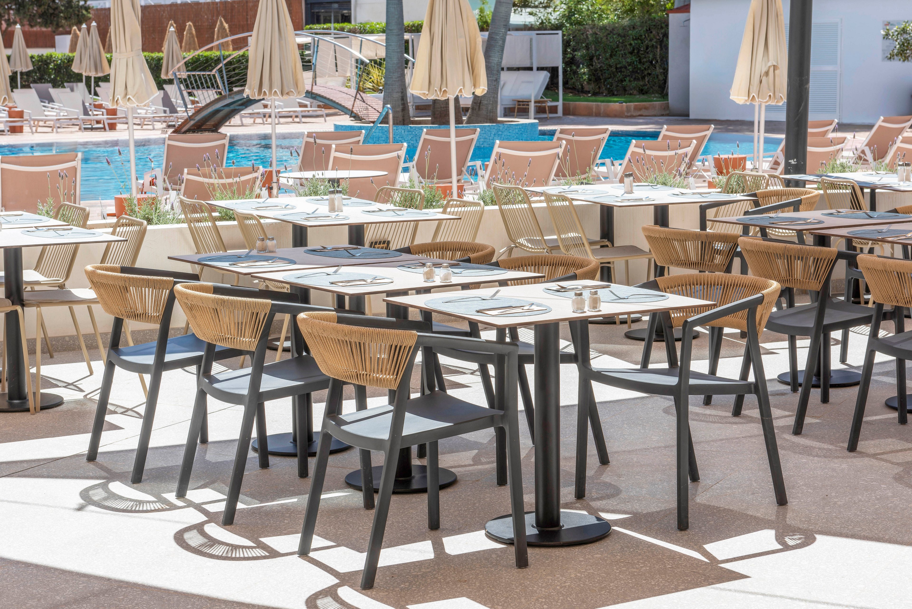 An outdoor restaurant terrace features neatly arranged tables and chairs beside a swimming pool. The modern seating includes woven chairs and simple black tables set with placemats and cutlery. Beige umbrellas provide shade over the dining area, while sun loungers line the pool in the background. The bright daylight and clean design create a relaxed, resort-like atmosphere.
