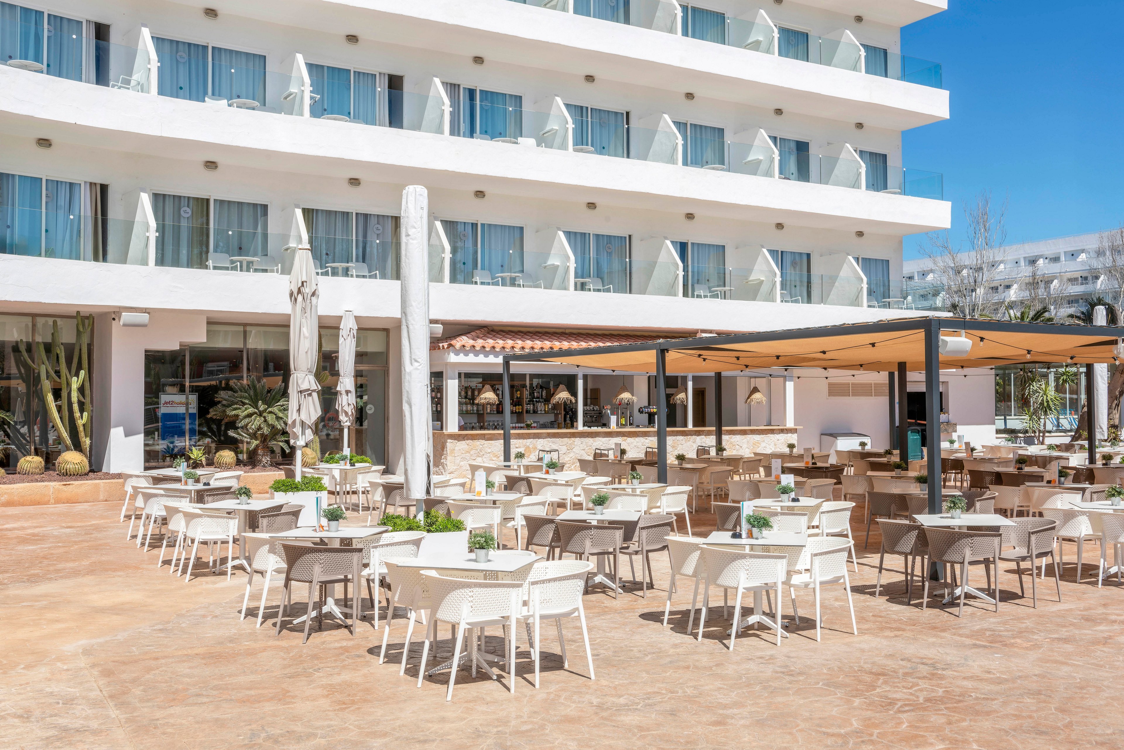 Spacious outdoor seating is arranged on a sunny terrace in front of a modern multi-story hotel. White tables and chairs are neatly organized under open sky and shaded pergolas. Closed umbrellas and potted plants add detail to the bright, minimalist courtyard. The scene suggests a relaxed resort atmosphere ready for guests.