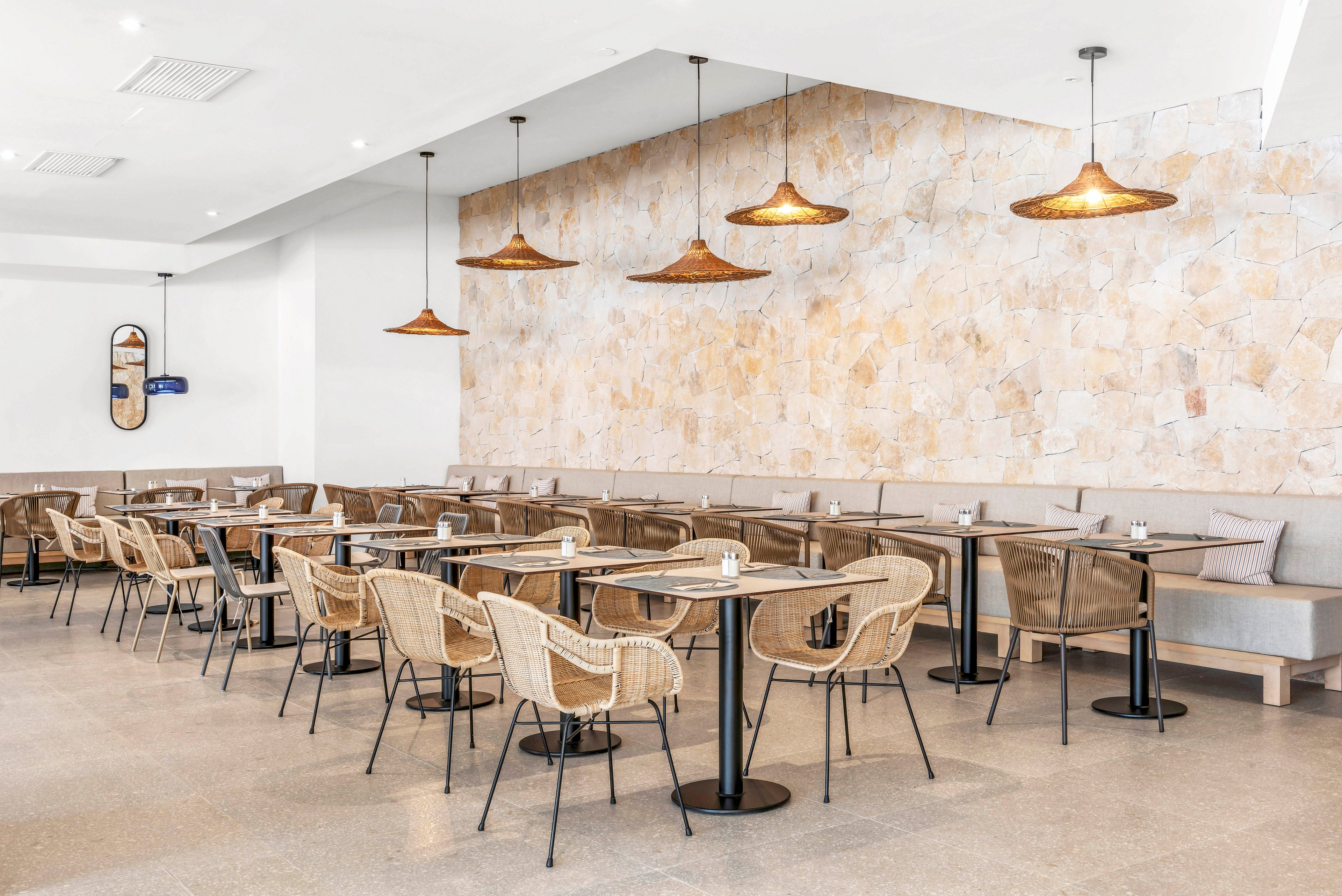 Spacious modern cafe interior featuring rows of tables with wicker chairs and bench seating. The room has a light stone accent wall and neutral flooring that enhance the airy atmosphere. Woven pendant lights hang from the ceiling, adding warmth and texture to the minimalist design. The space appears clean, empty, and ready to welcome guests.