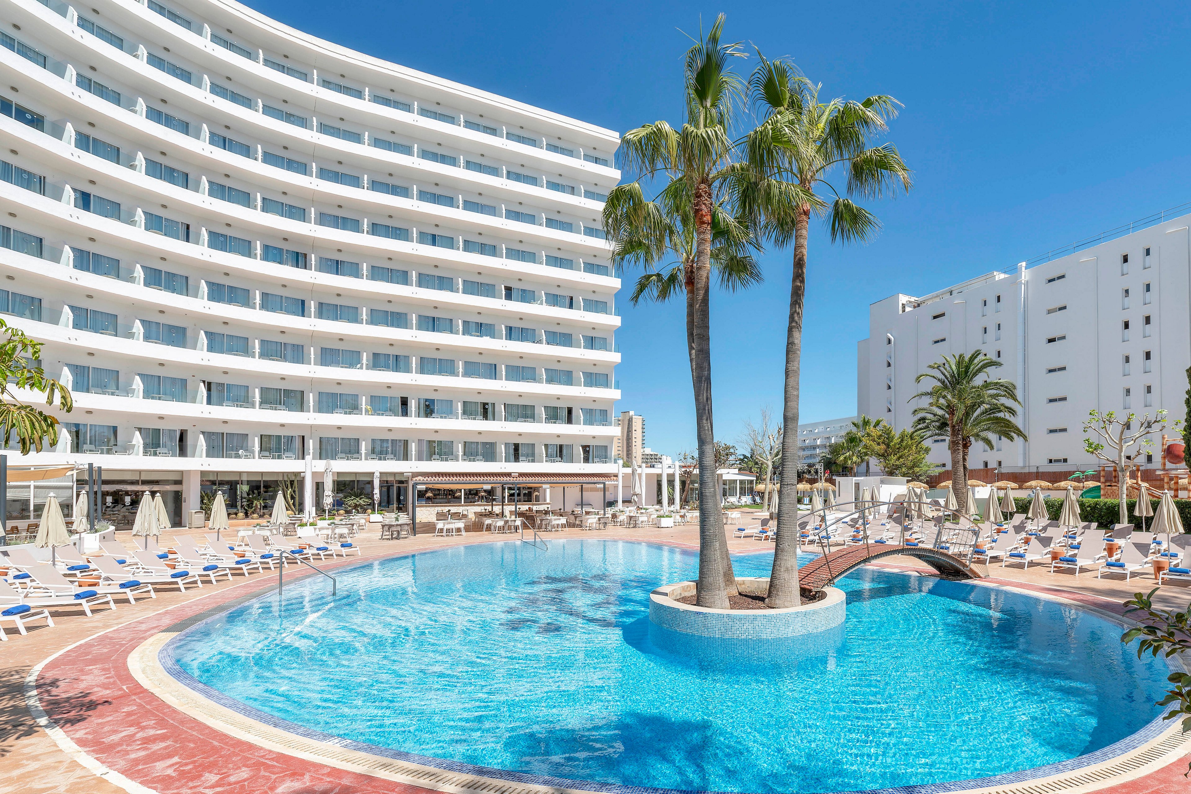 A large modern resort hotel curves around a bright blue outdoor swimming pool under a clear sky. Sun loungers and umbrellas line the poolside terrace, suggesting a relaxing vacation setting. Tall palm trees rise from an island in the center of the pool, adding a tropical touch. The scene is calm and inviting, with no visible guests, emphasizing the architecture and leisure facilities.
