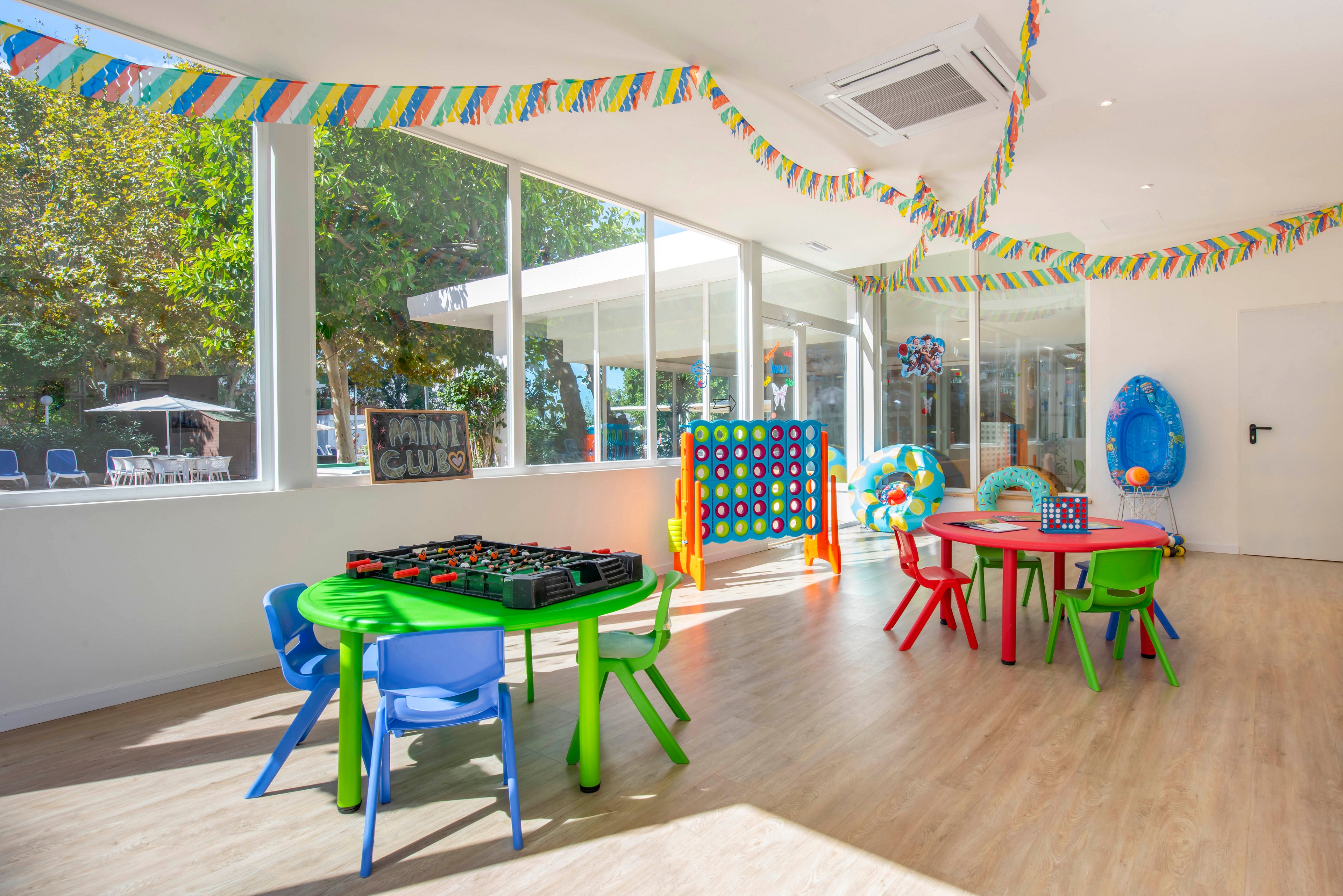 Colorful children’s tables and chairs are arranged in a bright indoor playroom. Large windows let in natural light and reveal trees and an outdoor area. Party streamers hang from the ceiling, and various games and inflatable toys are set up around the room. The space appears ready for kids’ activities or a birthday celebration.