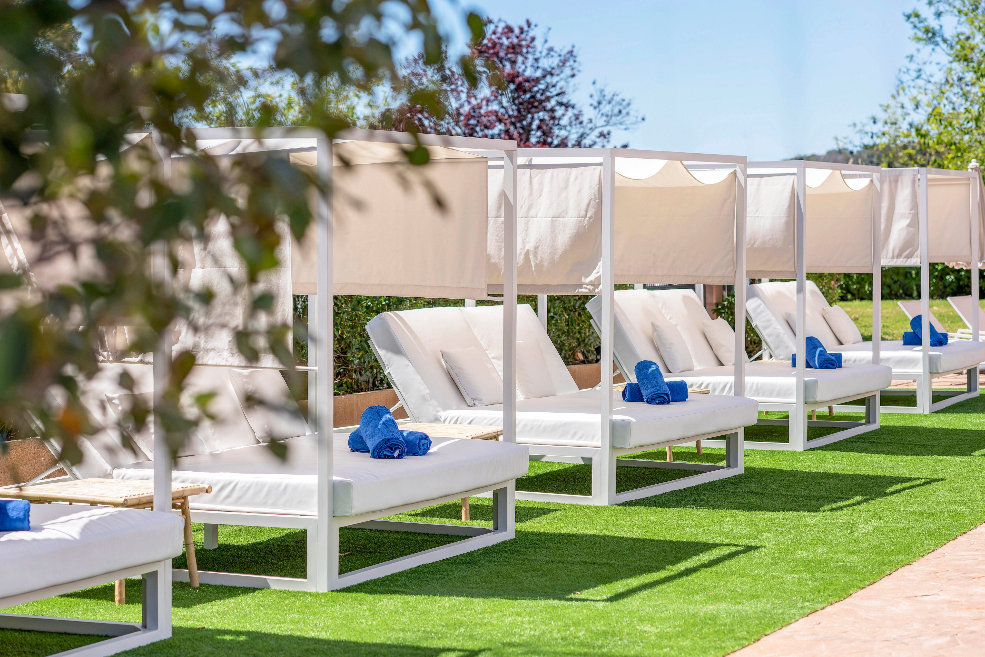 A row of white cabana daybeds is arranged neatly along a strip of bright green artificial grass. Each lounge bed is topped with plush white cushions and neatly rolled blue towels. Sunshades provide partial cover, suggesting a relaxing resort or hotel poolside setting. Trees and clear blue sky in the background enhance the calm, sunny atmosphere.