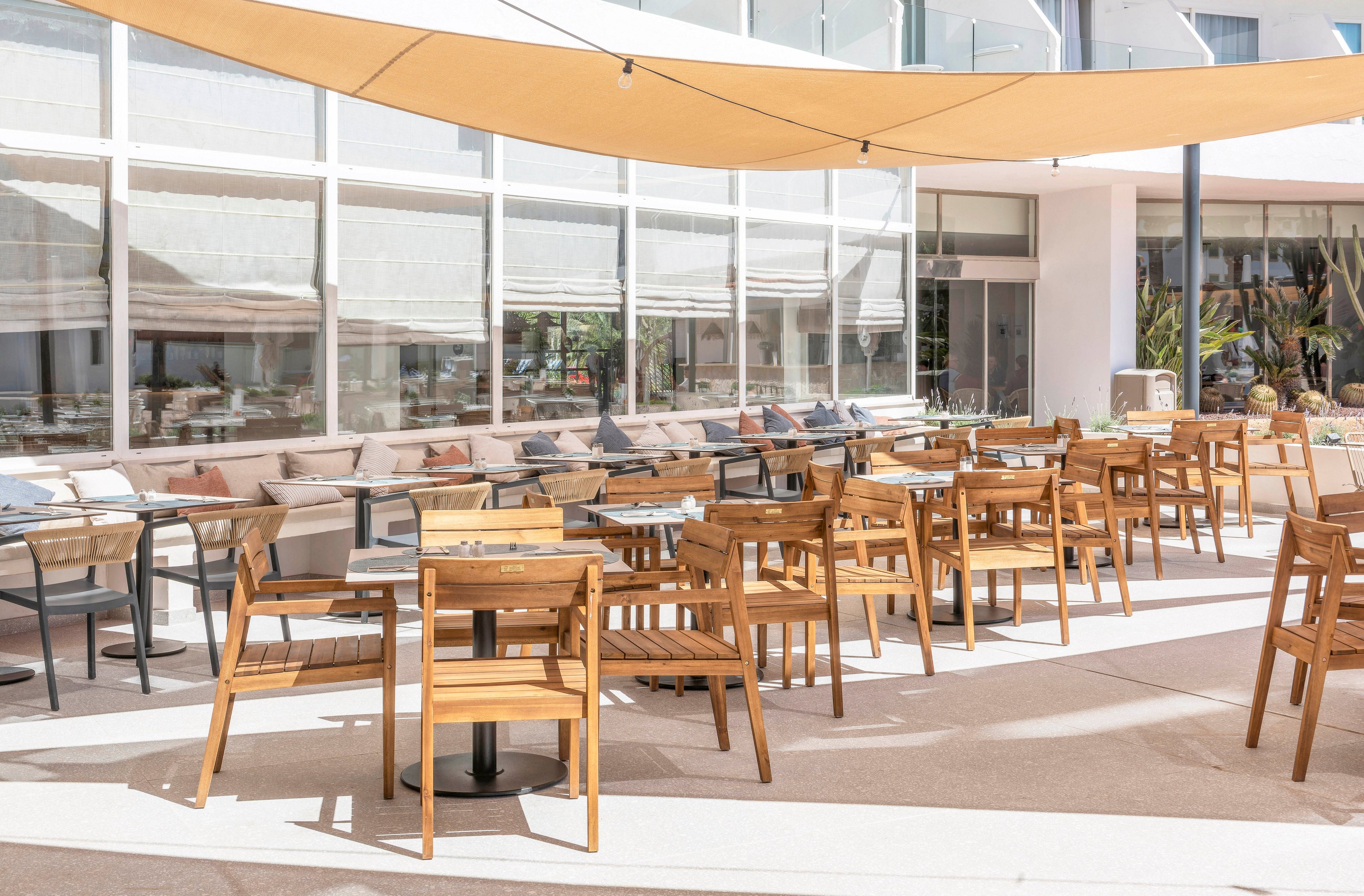 An outdoor cafe terrace features multiple wooden tables and chairs arranged neatly on a bright patio. Long cushioned bench seating lines the glass-walled building, creating a comfortable lounge area. Large fabric shade sails stretch overhead, casting soft shadows across the space. The overall scene suggests a modern, relaxed dining environment in warm, sunny weather.