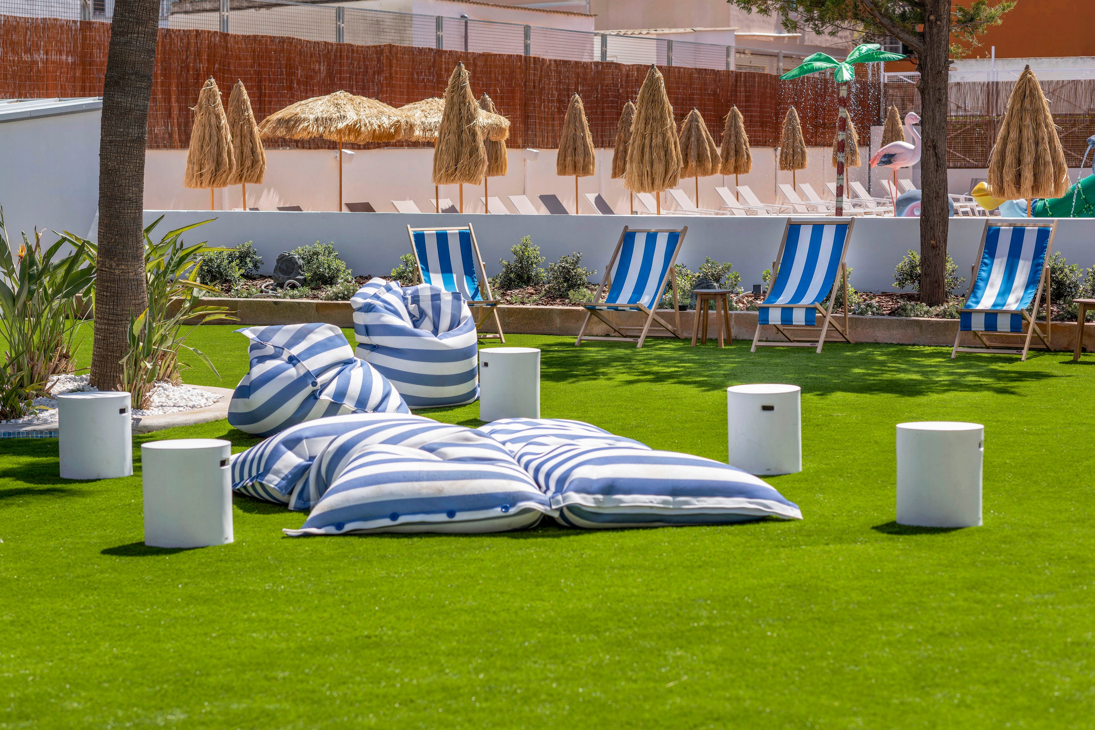 An outdoor lounge area features large blue and white striped floor cushions on a neatly manicured green lawn. Several matching striped deck chairs are arranged in the background near white walls and landscaped plants. Straw parasols provide shade, and small white cylindrical stools are placed around the seating area. The scene suggests a relaxed, resort-like atmosphere on a sunny day.