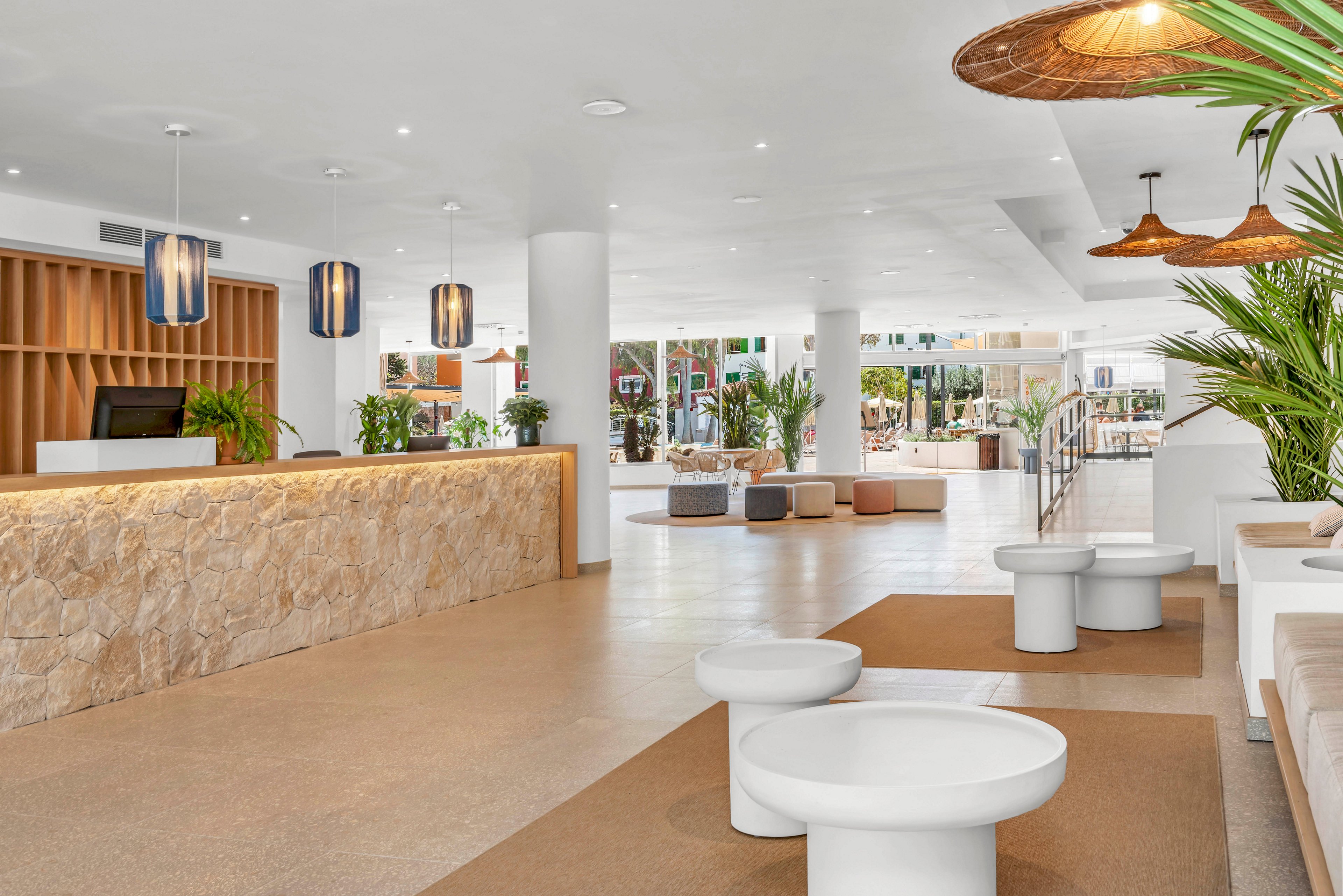 Spacious contemporary hotel lobby featuring a long stone reception desk on the left. The interior is bright and open with white walls, large columns, and minimalist furniture. Pendant lights and woven ceiling lamps add warmth to the clean design. Green plants and seating areas in the background create a welcoming atmosphere for guests.