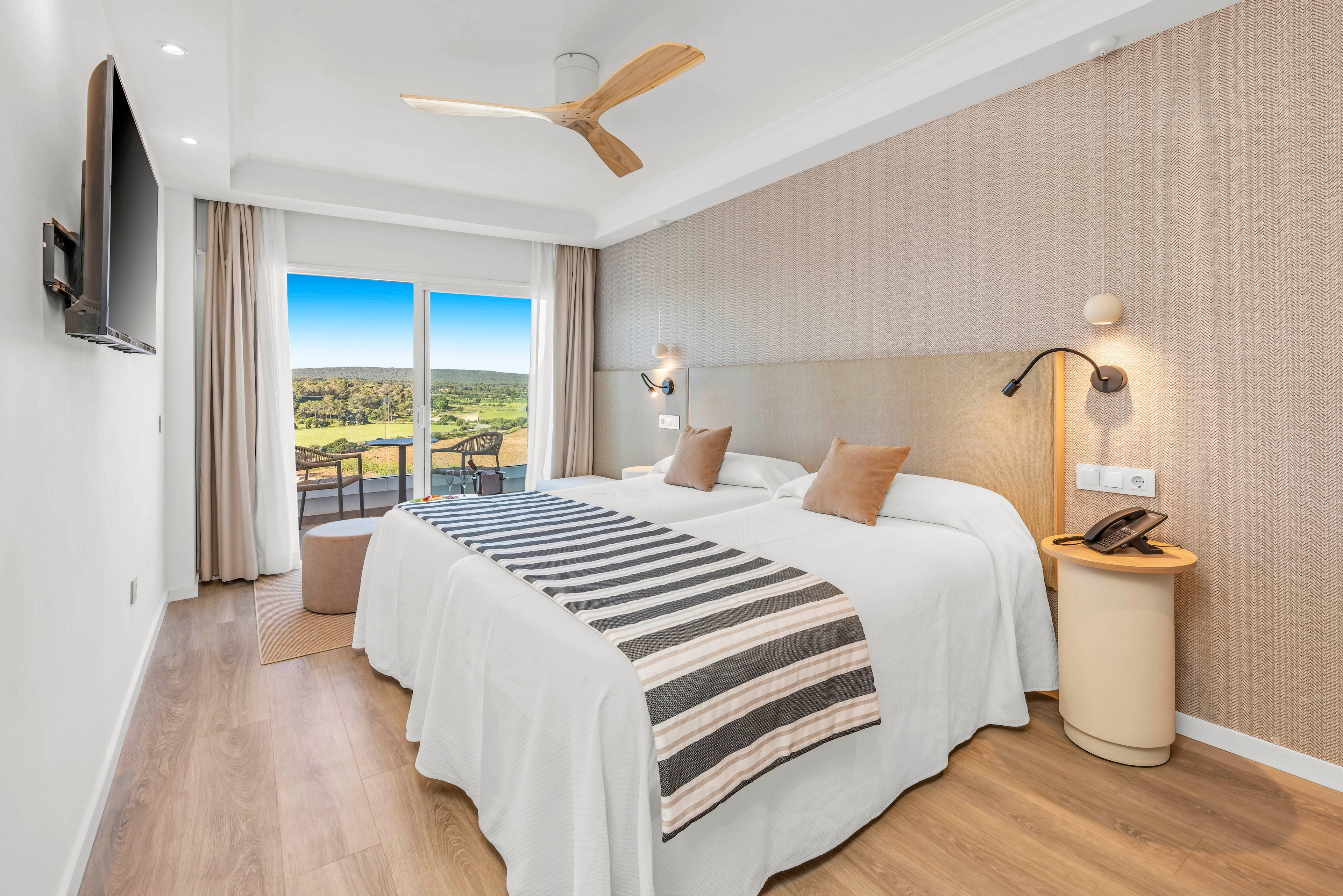 A bright modern hotel room features two beds pushed together with white linens and a striped throw. Warm wood flooring and neutral beige walls create a calm, minimalist atmosphere. A large sliding glass door opens to a balcony with chairs overlooking a green landscape under a blue sky. Contemporary lighting, a ceiling fan, and a wall-mounted television complete the comfortable interior.