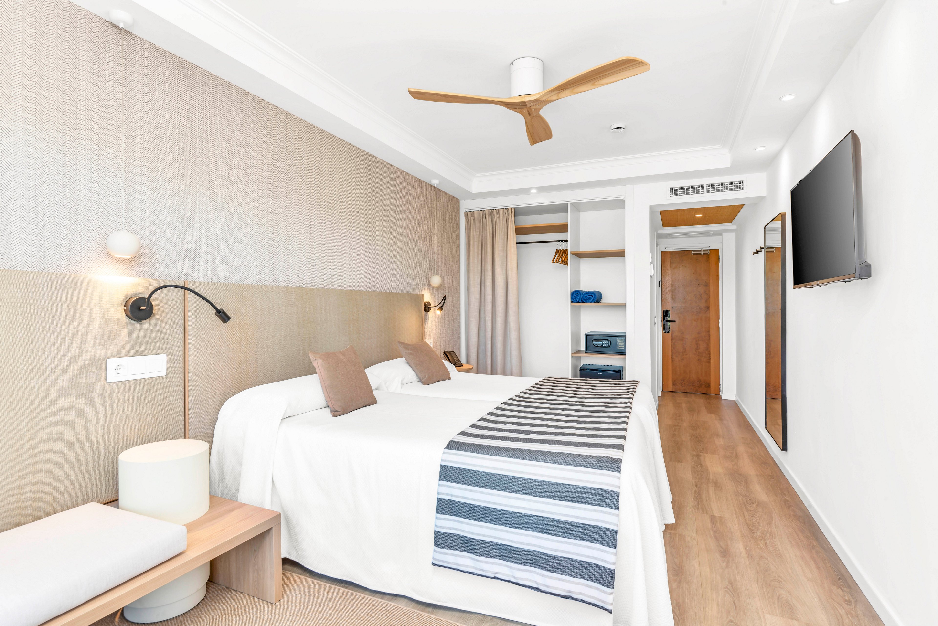 A bright modern hotel bedroom features a large neatly made bed with white linens and a striped runner. The room includes built-in open shelving, a wall-mounted television, and a wooden entrance door. Soft neutral tones, wood flooring, and a contemporary ceiling fan create a clean, minimalist atmosphere. Recessed lighting and natural light enhance the airy, welcoming feel of the space.