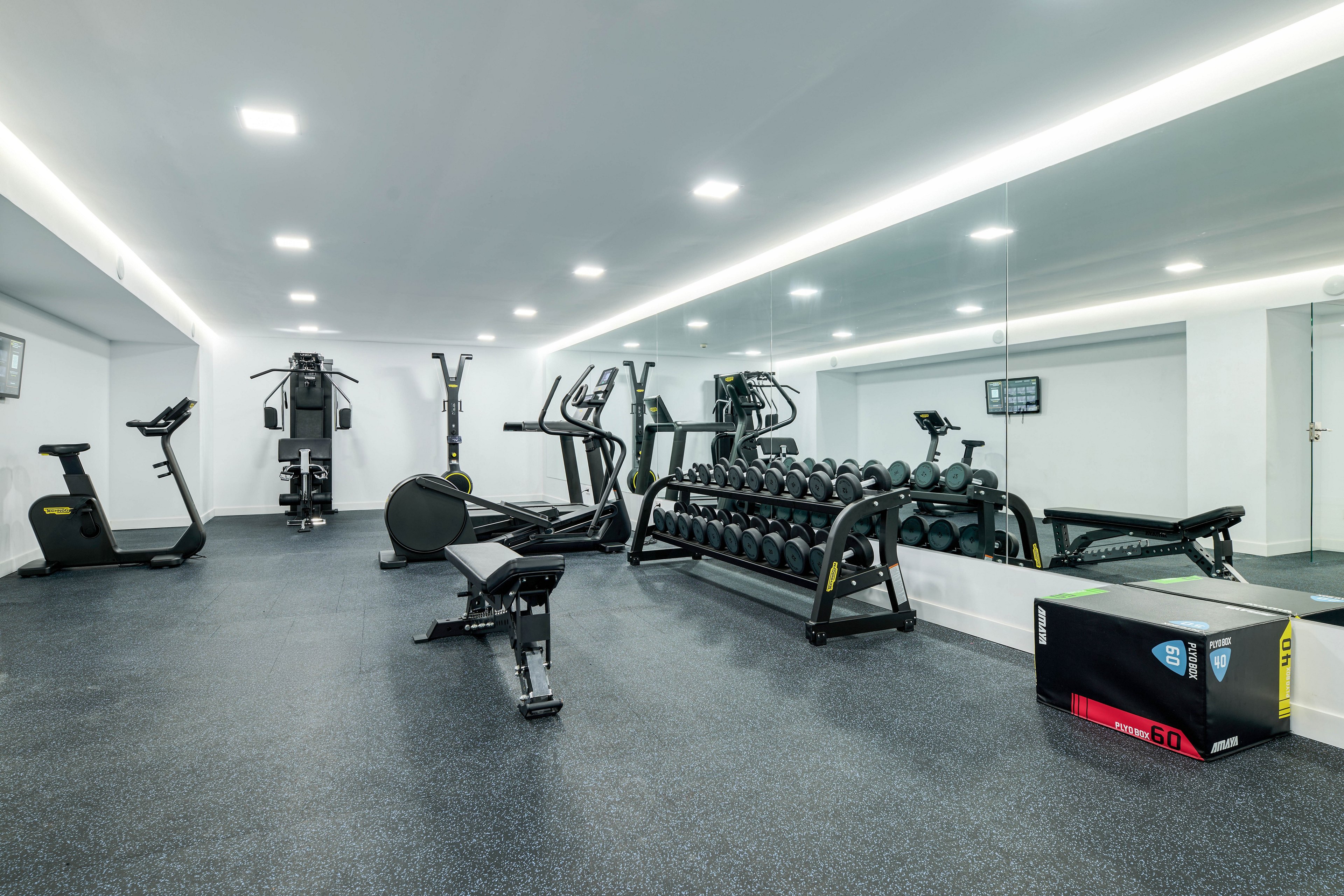 A clean, modern gym interior features rows of dumbbells and various strength-training machines. The space is empty, with a bench and cardio equipment arranged on a dark rubber floor. A large wall mirror reflects the equipment and bright ceiling lights, enhancing the sense of space. A black and red exercise box with the number 60 printed on it sits in the foreground.