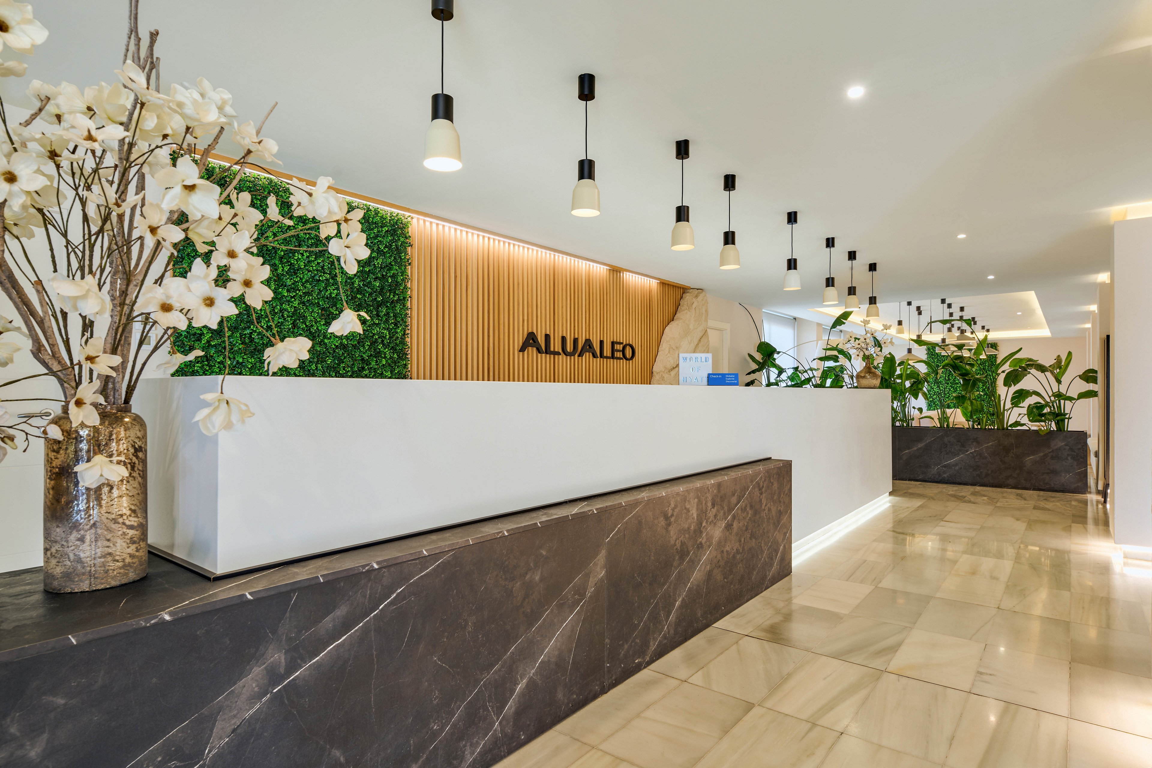 A contemporary hotel reception desk features a long white counter set against a wooden slat wall with green plant panels. The word "ALUA LEO" is prominently displayed on the back wall behind the desk. Pendant lights hang from the ceiling, illuminating the polished floor and sleek dark stone surfaces. Decorative white flowers and lush indoor plants add a fresh, welcoming atmosphere to the lobby space.
