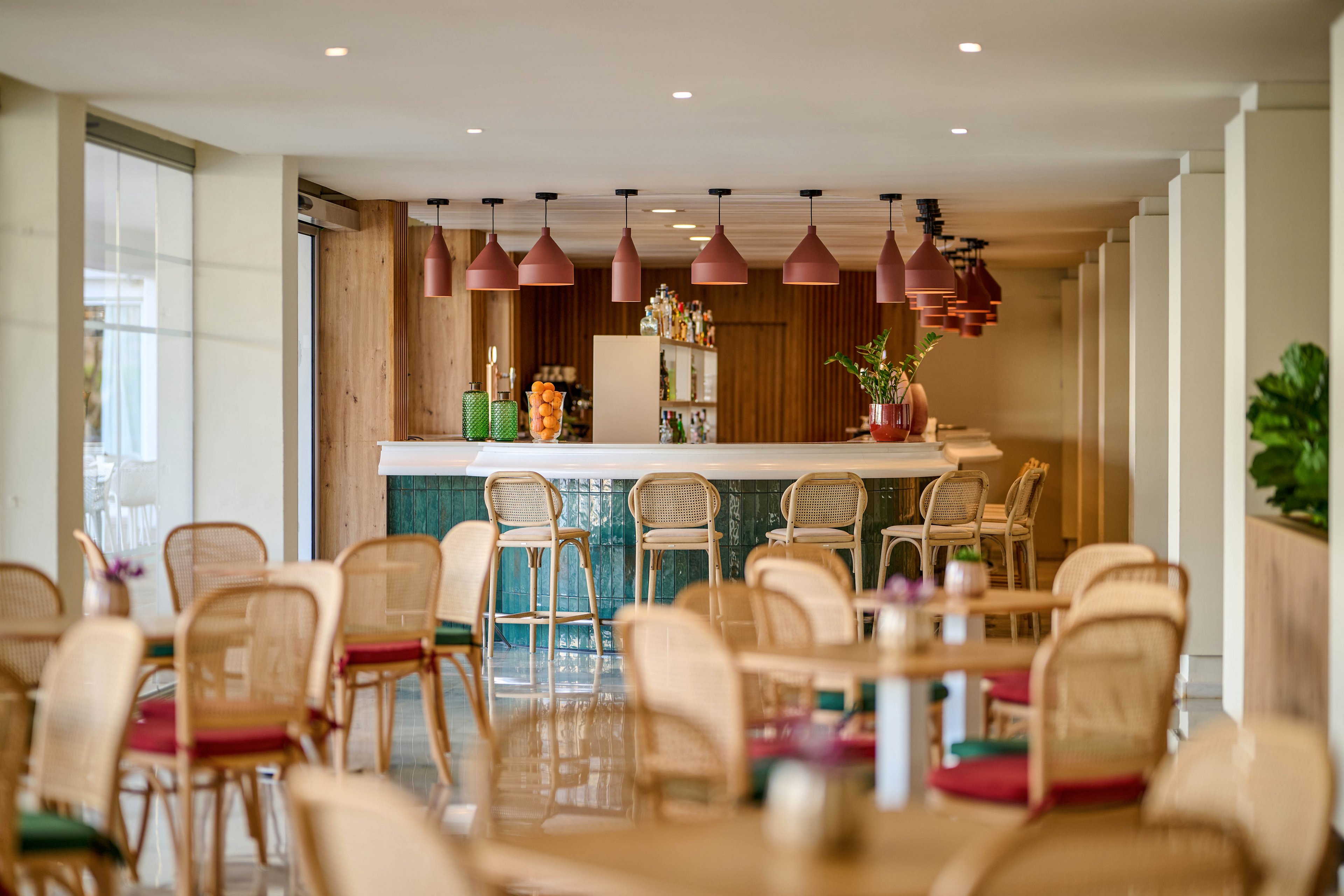 A contemporary bar area features a long counter with a green tiled front and a white countertop. Wicker chairs and tables fill the foreground, creating a relaxed lounge atmosphere. Pink pendant lights hang in a row above the bar, illuminating bottles and glassware on the shelves. Large windows and neutral walls give the space an airy, bright feel with subtle greenery accents.