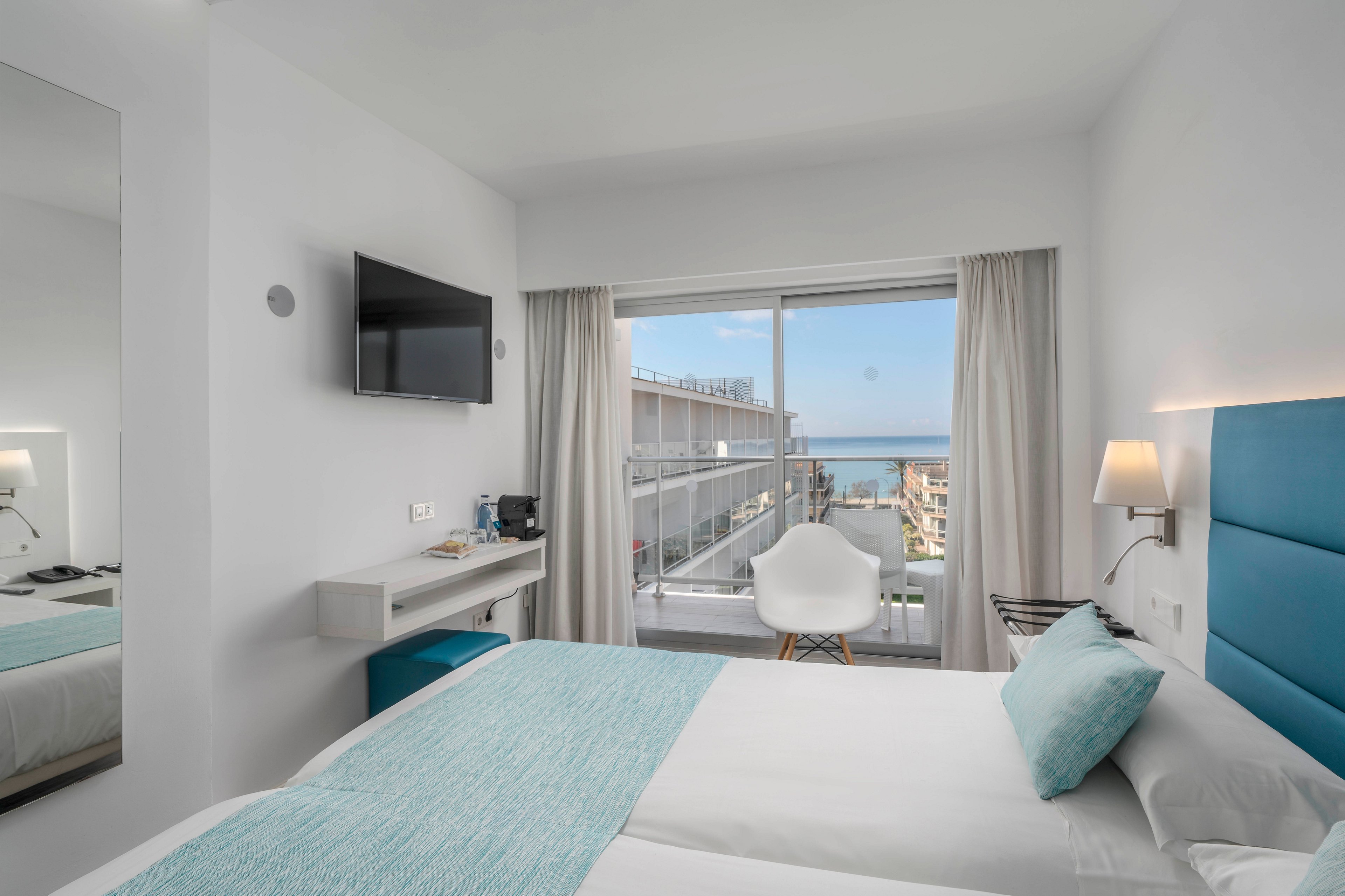 Bright modern hotel room features a neatly made double bed with turquoise accents. A wall-mounted television and compact desk area sit opposite the bed, with a coffee station and chair nearby. Large sliding glass doors open to a balcony overlooking nearby buildings and the distant sea under a blue sky. Soft neutral walls, white curtains, and minimal decor create a clean, contemporary atmosphere.