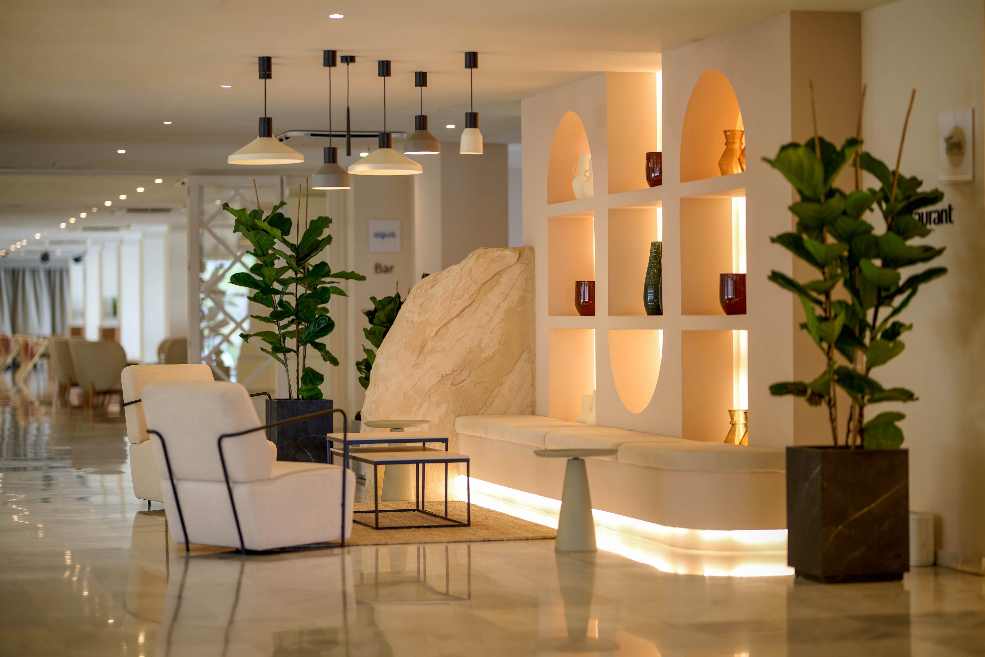 A contemporary hotel lobby features white lounge chairs and a small metal-framed table arranged on a glossy marble floor. Built-in illuminated shelves display decorative vases and objects, creating a warm focal point. Tall potted plants flank the seating area, adding greenery to the neutral interior. Pendant lights hang from the ceiling, enhancing the soft, inviting ambiance of the modern space.