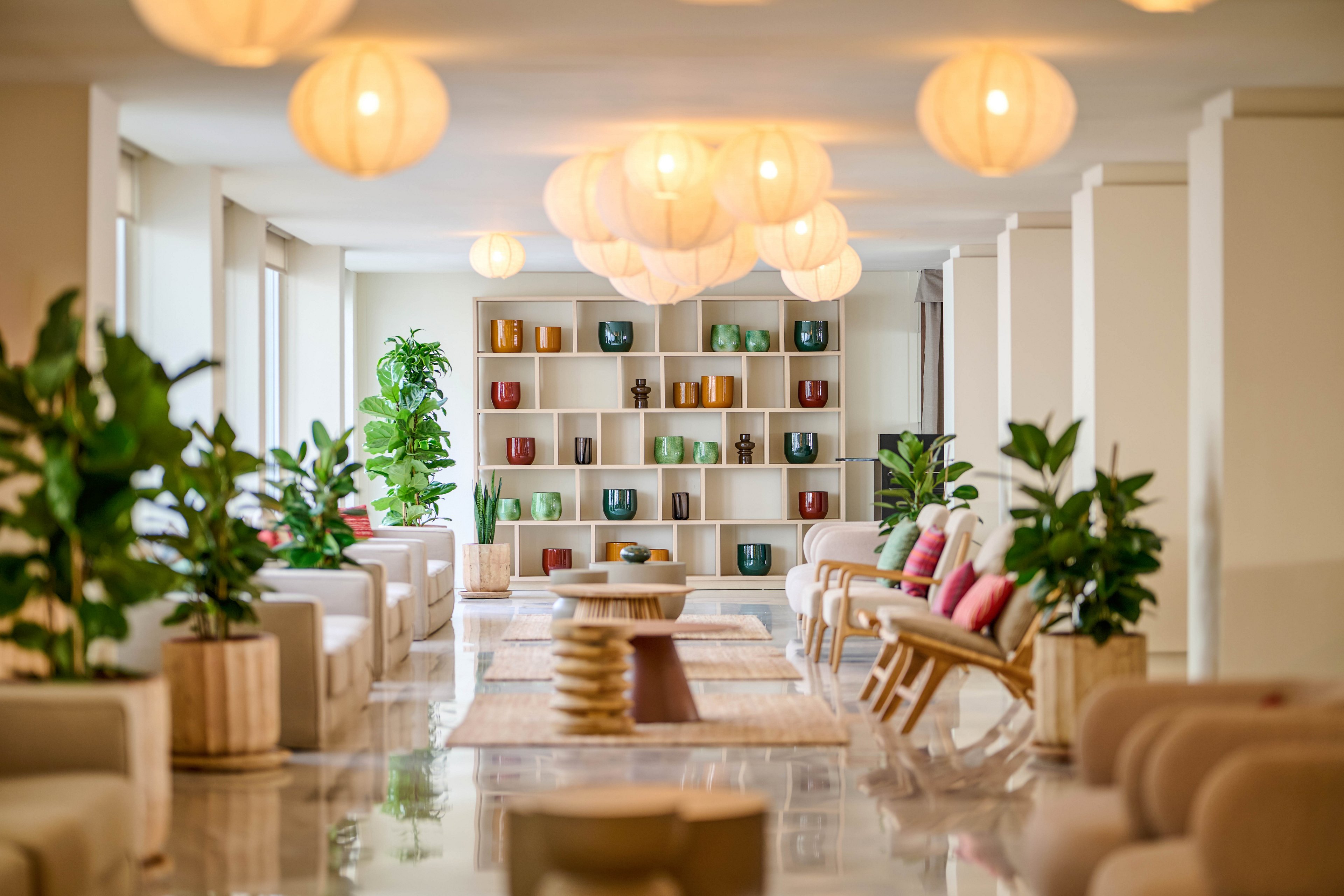 A spacious modern lounge features low seating arranged along a glossy floor. Large potted plants and neutral-toned furniture create a relaxed, contemporary atmosphere. Warm pendant lights hang from the ceiling, casting a soft glow across the room. At the far end, a shelving unit displays colorful decorative vases and objects, adding visual interest to the minimalist space.