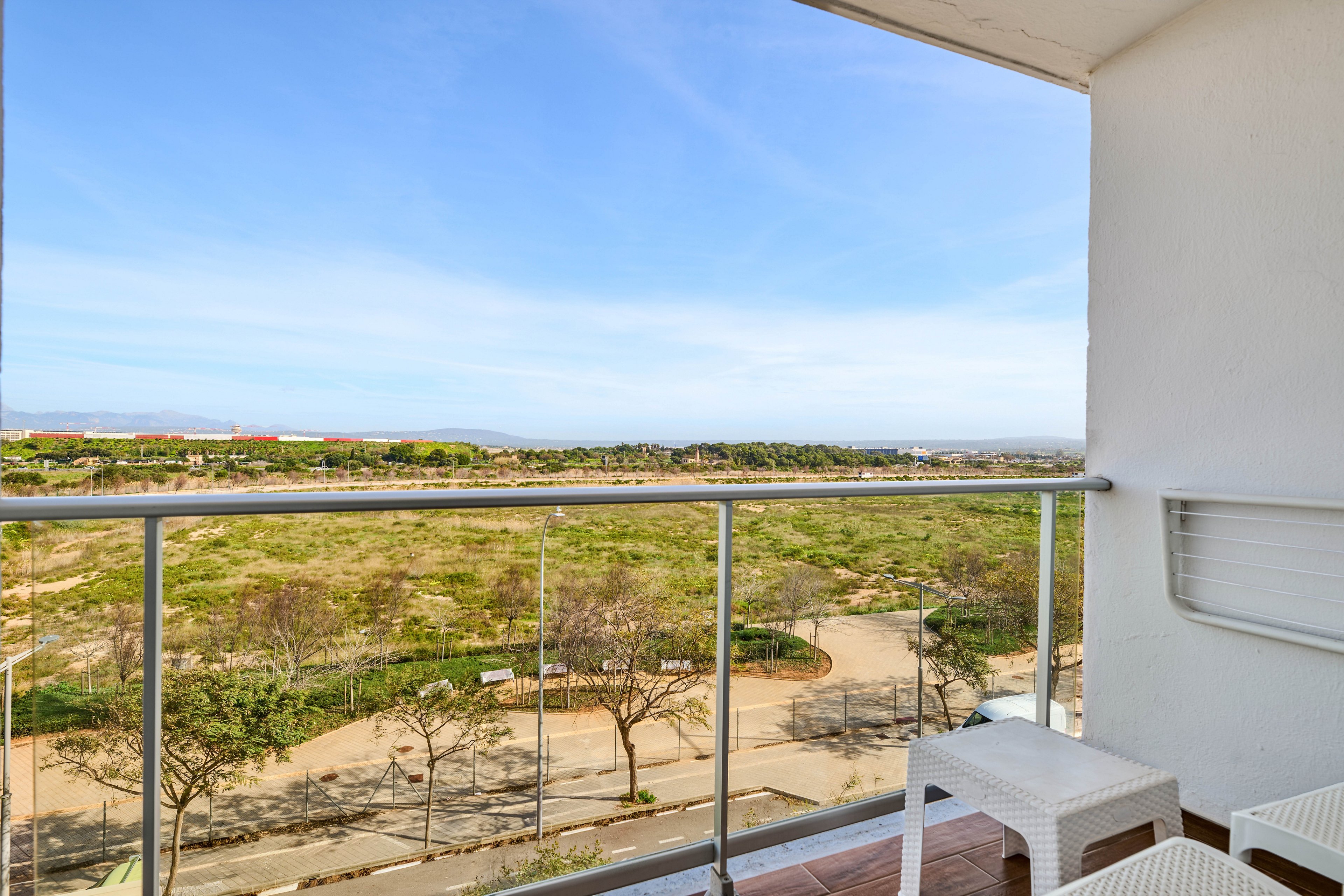 A small balcony with white outdoor furniture overlooks a green park and open landscape. Metal railings frame the view of trees, pathways, and a distant horizon under a clear blue sky. The scene suggests a residential apartment setting with a peaceful, elevated vantage point. Soft natural light and minimal decor create a calm, airy atmosphere.