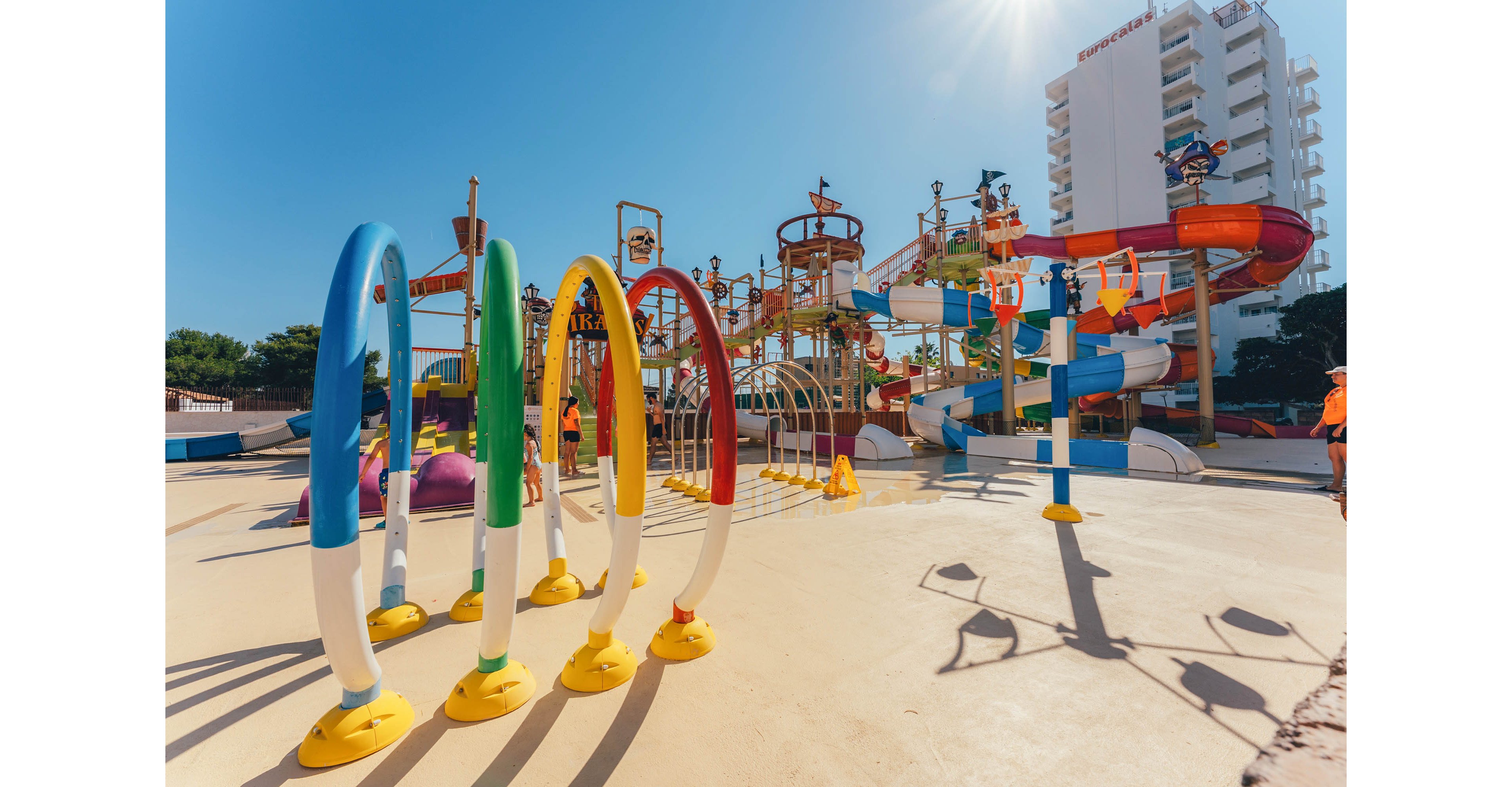 A vibrant outdoor water park features colorful hoops and play structures under a clear blue sky. Multiple slides, climbing towers, and splash elements create an exciting family-friendly environment. A few people in swimwear and life vests are visible near the attractions, suggesting supervised recreational activity. Tall buildings and trees in the background indicate a resort or urban vacation setting.