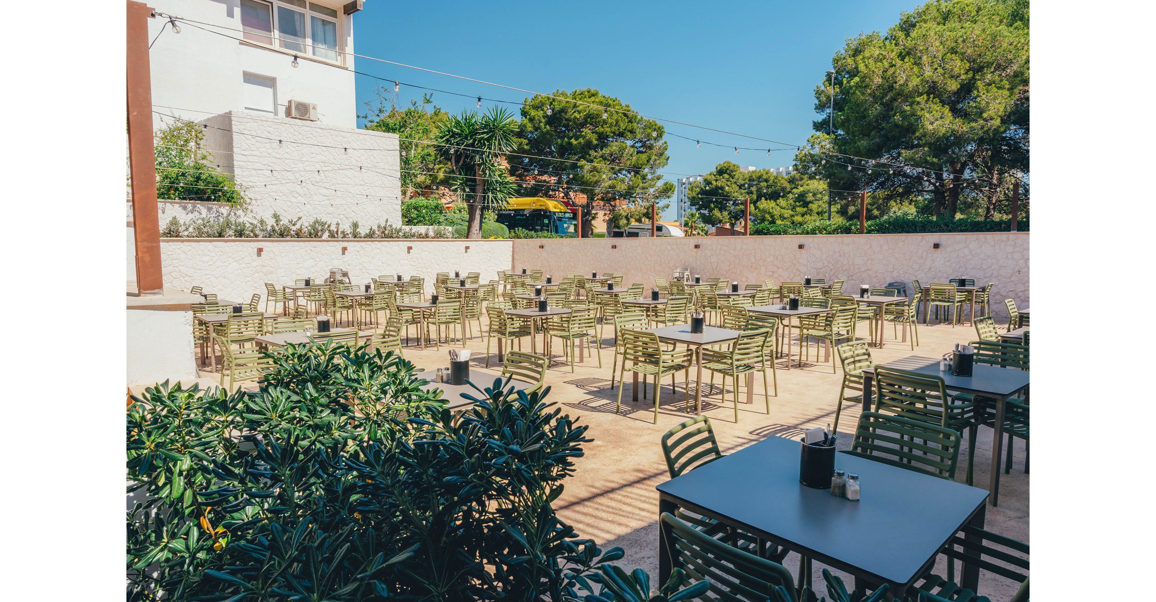 An open-air restaurant terrace is filled with neatly arranged green metal tables and chairs. The space is empty of people, suggesting preparation before guests arrive or a quiet time of day. String lights hang overhead, and lush trees and shrubs surround the patio, creating a relaxed atmosphere. A bright, clear sky and strong sunlight emphasize the warm, summery setting.