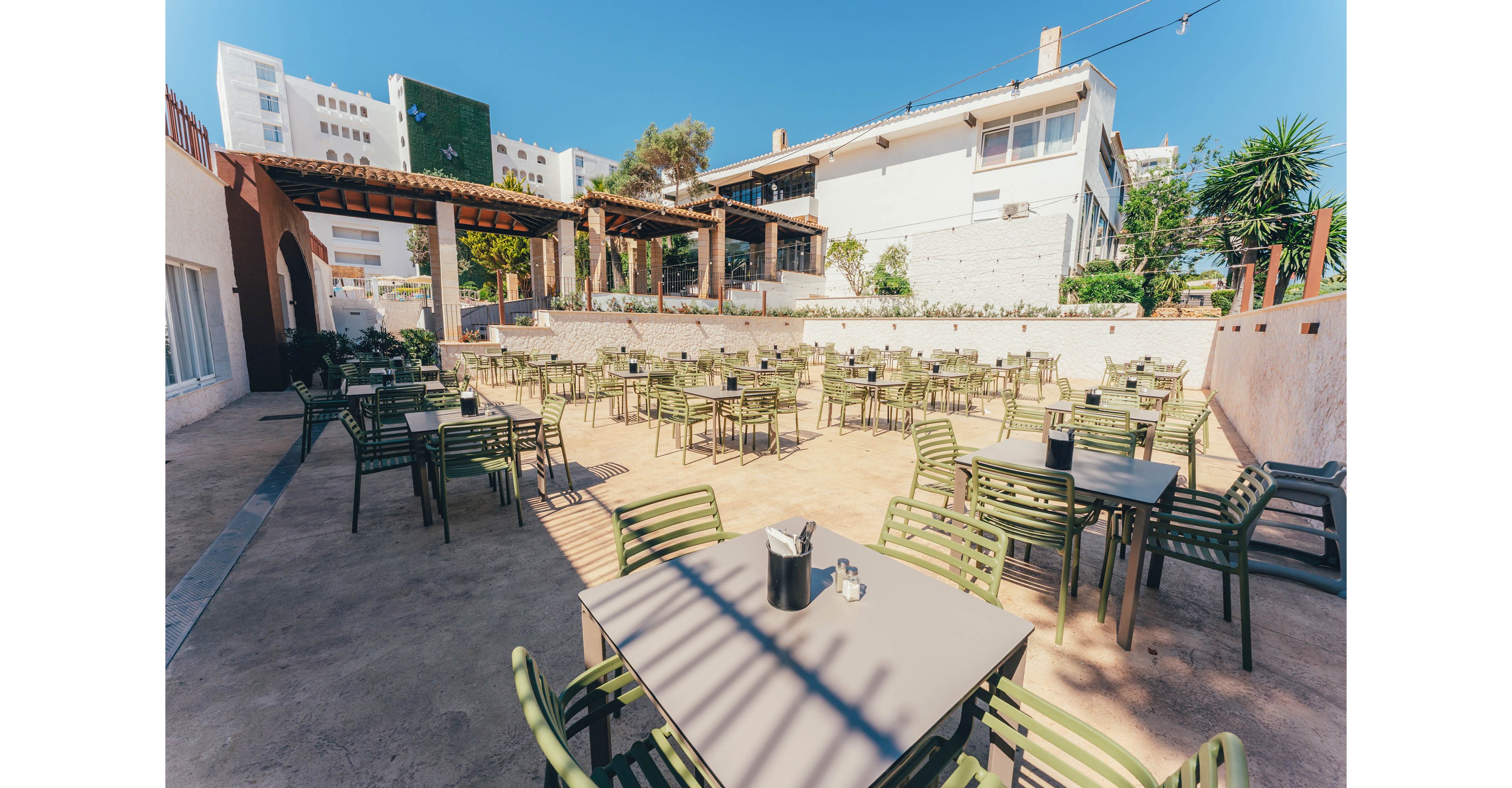 An empty outdoor cafe terrace is filled with neatly arranged tables and green metal chairs. The seating area is set in a sunny courtyard beside light-colored buildings and a shaded pergola. Bright midday light casts strong shadows across the ground, emphasizing the orderly layout. The scene feels calm and ready for guests, with clear blue sky overhead and some greenery around the perimeter.