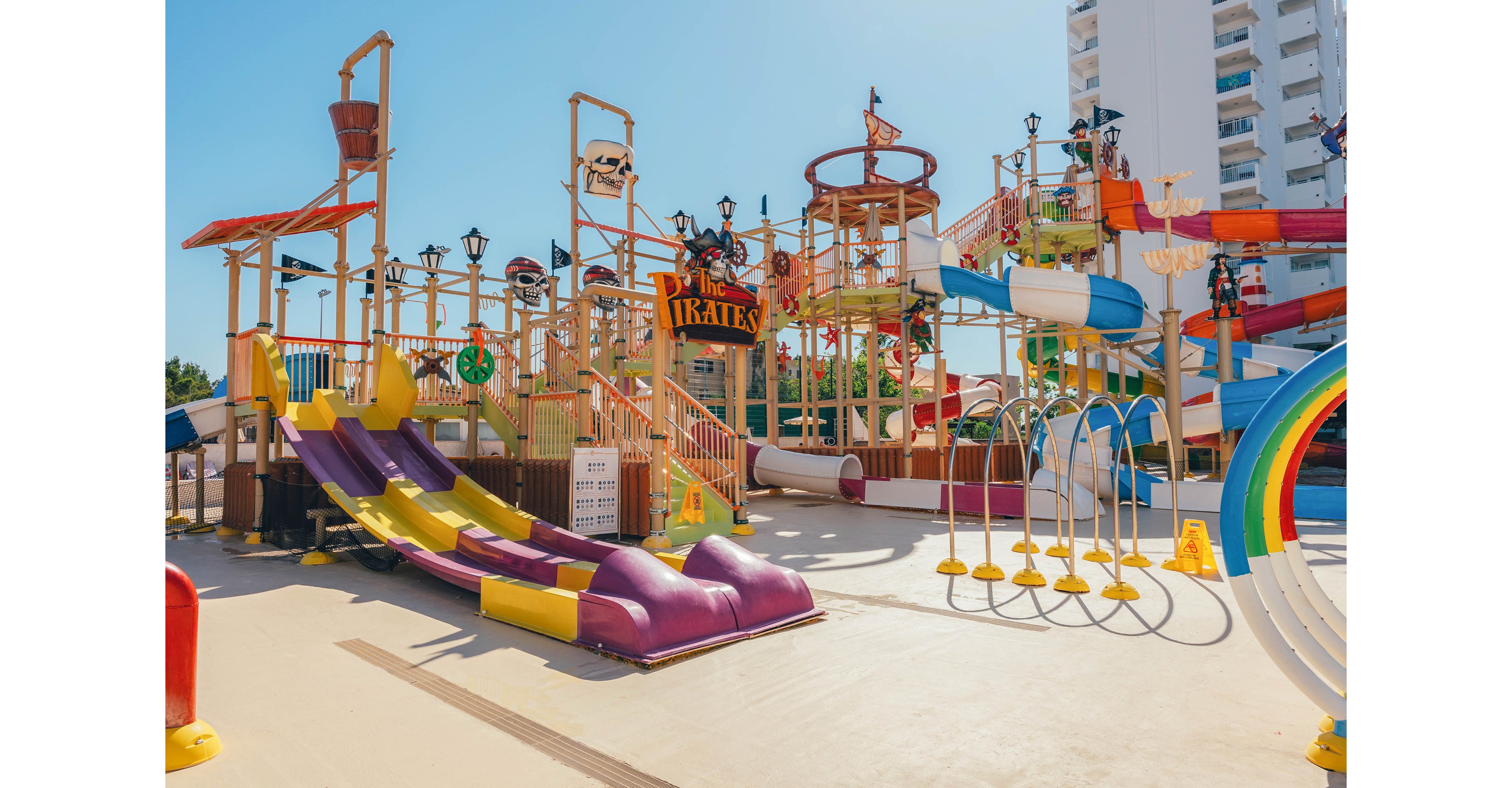 A vibrant pirate-themed water playground features multiple slides, climbing structures, and splash elements. Bright yellow, red, purple, and orange colors dominate the play area, creating a fun and inviting atmosphere. The central structure includes pirate decorations and a sign reading "PIRATES" above the stairs. In the background, a tall modern building rises behind the water park under a clear blue sky.