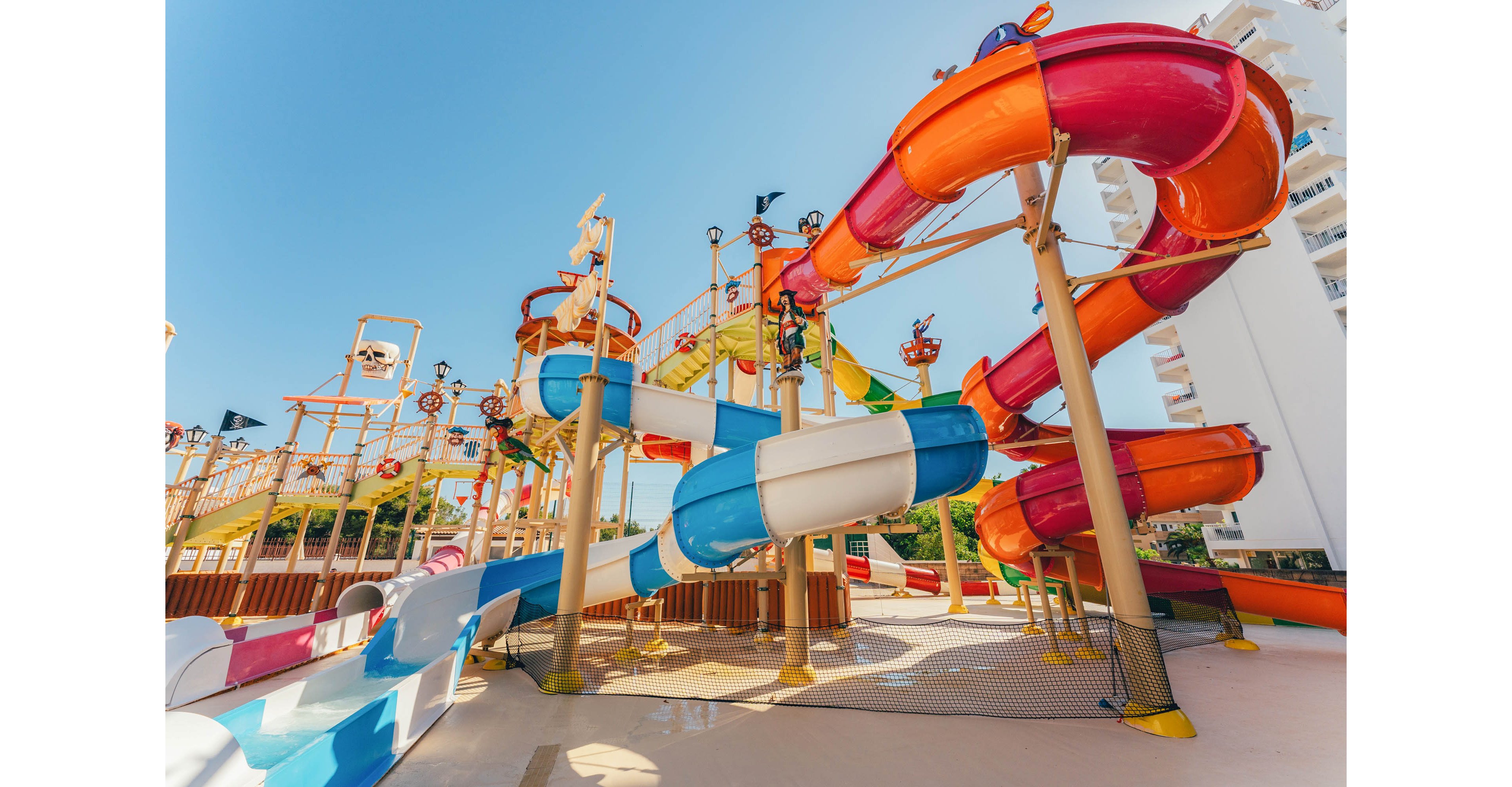 A vibrant outdoor water park features a network of twisting slides in bright orange, pink, blue, and white. The structure includes elevated platforms, stairways, and water play elements under a clear blue sky. No people are visible, emphasizing the architecture and colorful tubes. Nearby buildings frame the scene, suggesting a resort or vacation destination.