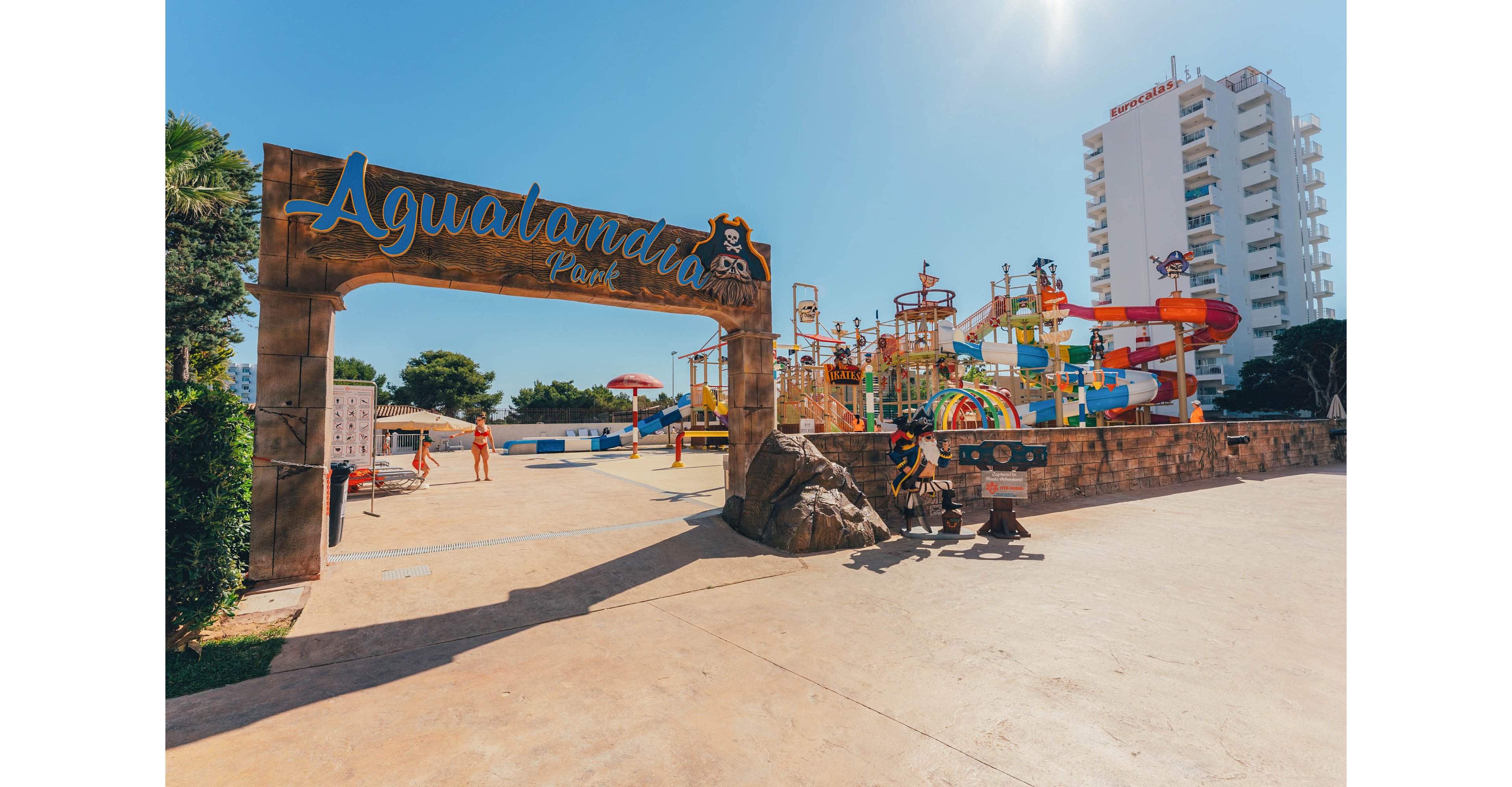 Wide view of the Aqualandia Park entrance leading into a colorful outdoor water playground. The scene shows slides, splash structures, and water features under a clear blue sky. A few people and children are visible enjoying the warm weather near the pools. A tall white hotel or apartment building rises in the background, emphasizing the resort setting.