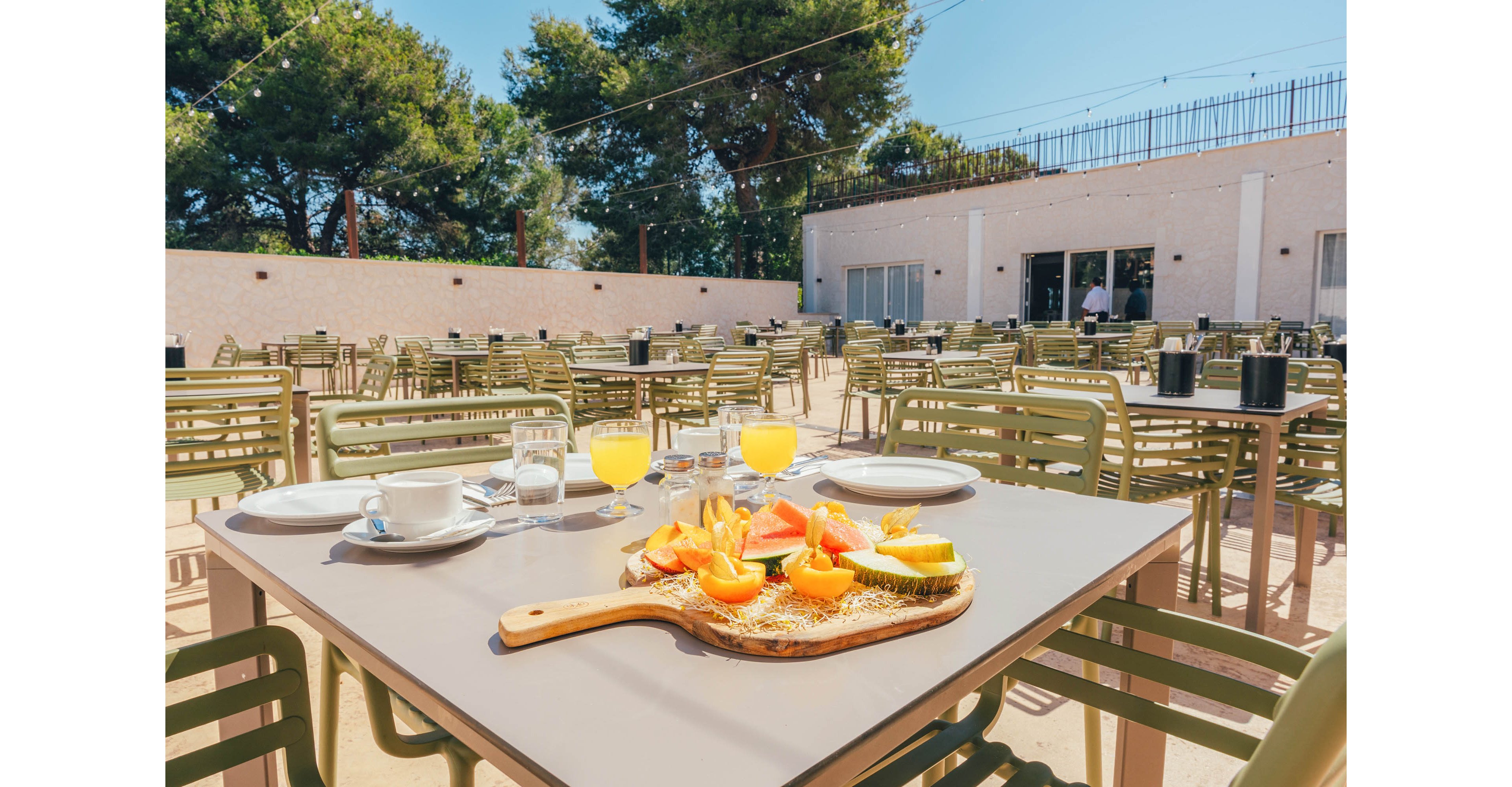 A wooden board with sliced fresh fruit and pastries is arranged on a cafe table outdoors. Two glasses of orange juice, a cup of coffee, plates, and cutlery are set for a meal. The terrace is filled with empty green chairs and tables under a clear sunny sky. Tall trees and a light-colored building frame the relaxed, summery setting.