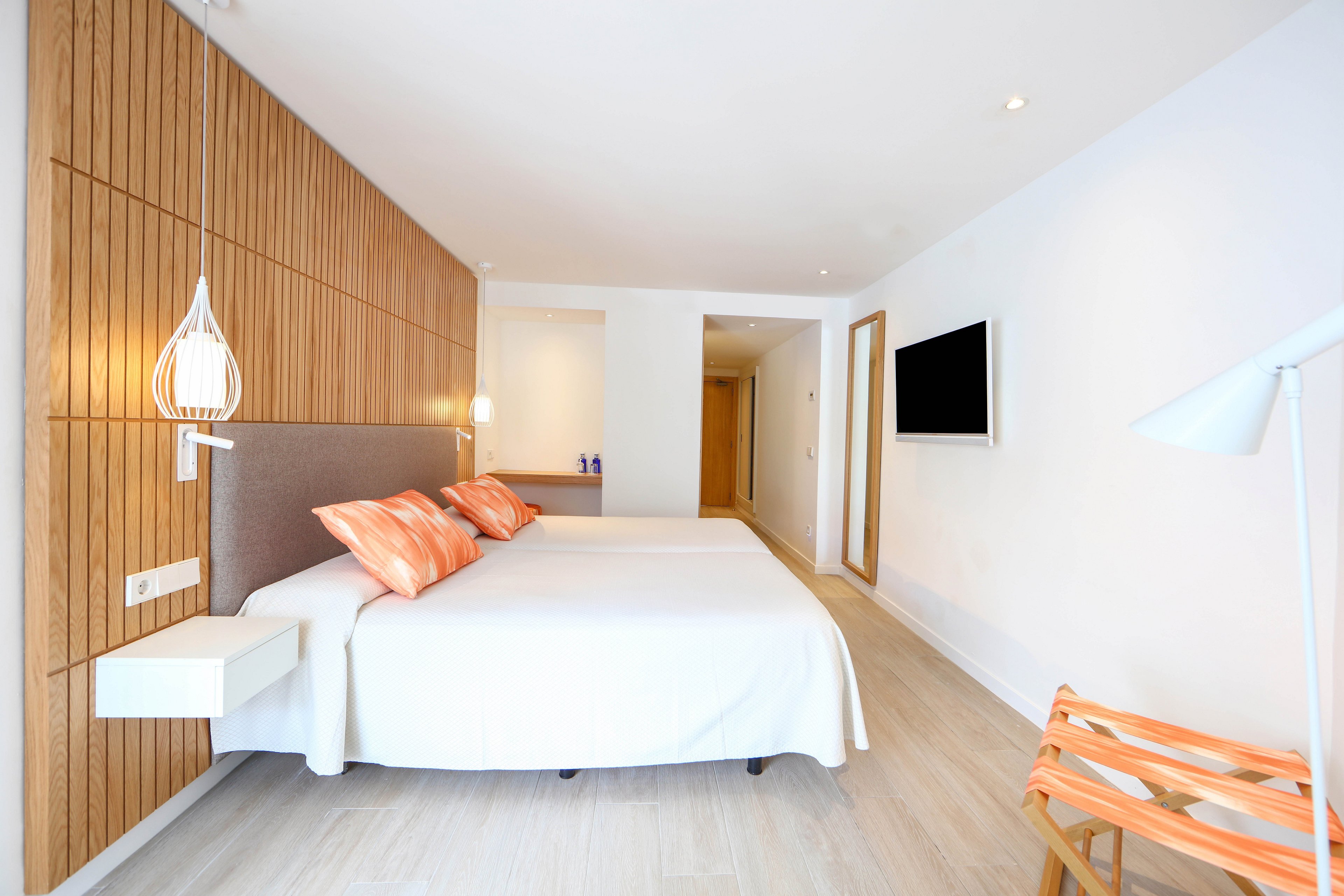 A contemporary hotel bedroom features a double bed with white linens and orange patterned pillows. The room has light wooden flooring and a vertical wood panel accent wall behind the bed. A flat-screen television is mounted on the white wall opposite the bed, and a minimalist white lamp and small bedside shelf add to the clean design. The space is brightly lit, creating an airy and inviting atmosphere with a touch of coastal style.
