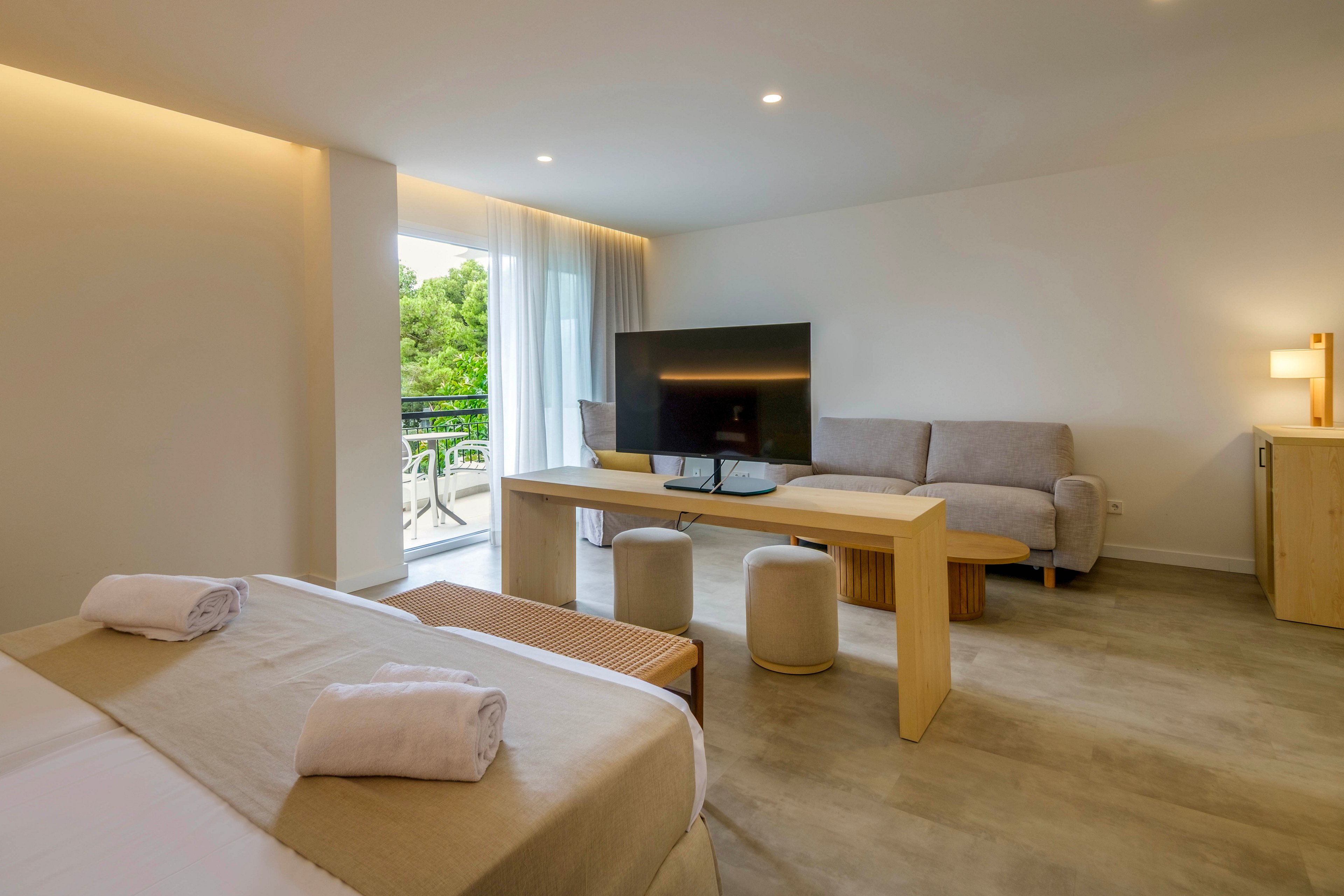 A spacious modern hotel room features a large bed in the foreground with neatly rolled white towels. A wooden table supports a flat-screen television facing a light gray sofa and small cushioned stools. Floor-to-ceiling windows with sheer curtains open to a balcony overlooking green trees. Warm recessed lighting and neutral tones create a calm, minimalist atmosphere.