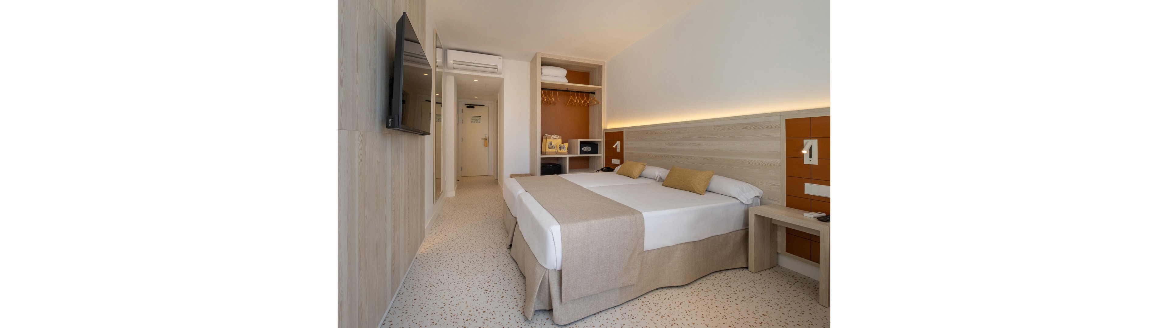 A contemporary hotel room features a neatly made double bed with neutral-toned bedding and accent pillows. Built-in open closets with shelves and hanging space are visible near the entrance, along with a small desk and bedside tables. Warm indirect lighting runs along the headboard, complementing the light wood finishes and speckled flooring. A flat-screen television is mounted on the wall opposite the bed, creating a clean and functional guest space.