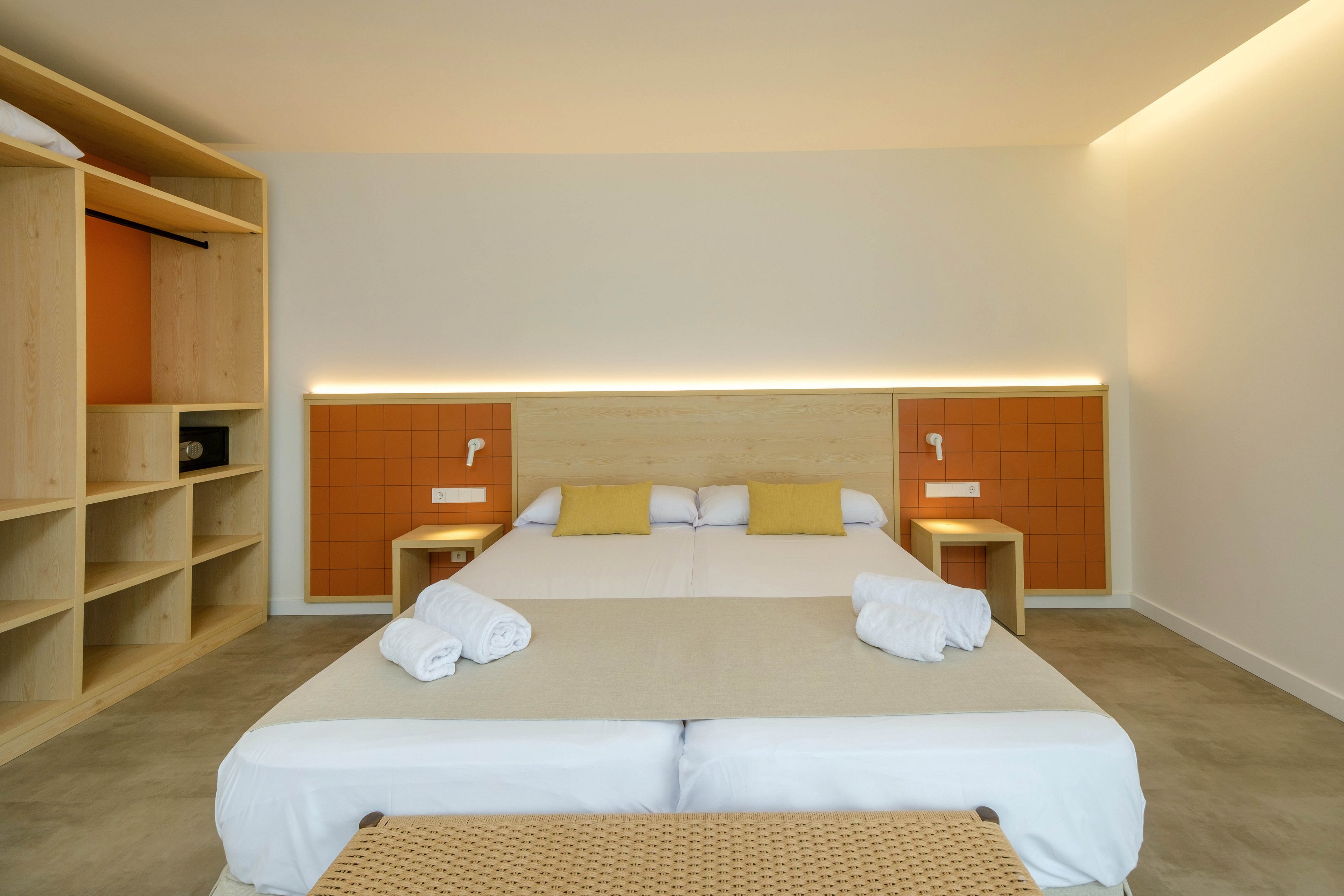 A modern hotel bedroom features a large double bed with a light wood headboard and integrated lighting. Two bedside tables with mounted reading lamps flank the bed, and neatly folded white towels rest on the foot of the bed. Open shelving and storage units line one side of the room, adding to the functional design. The overall space feels bright and minimalist with neutral tones and subtle orange accents.