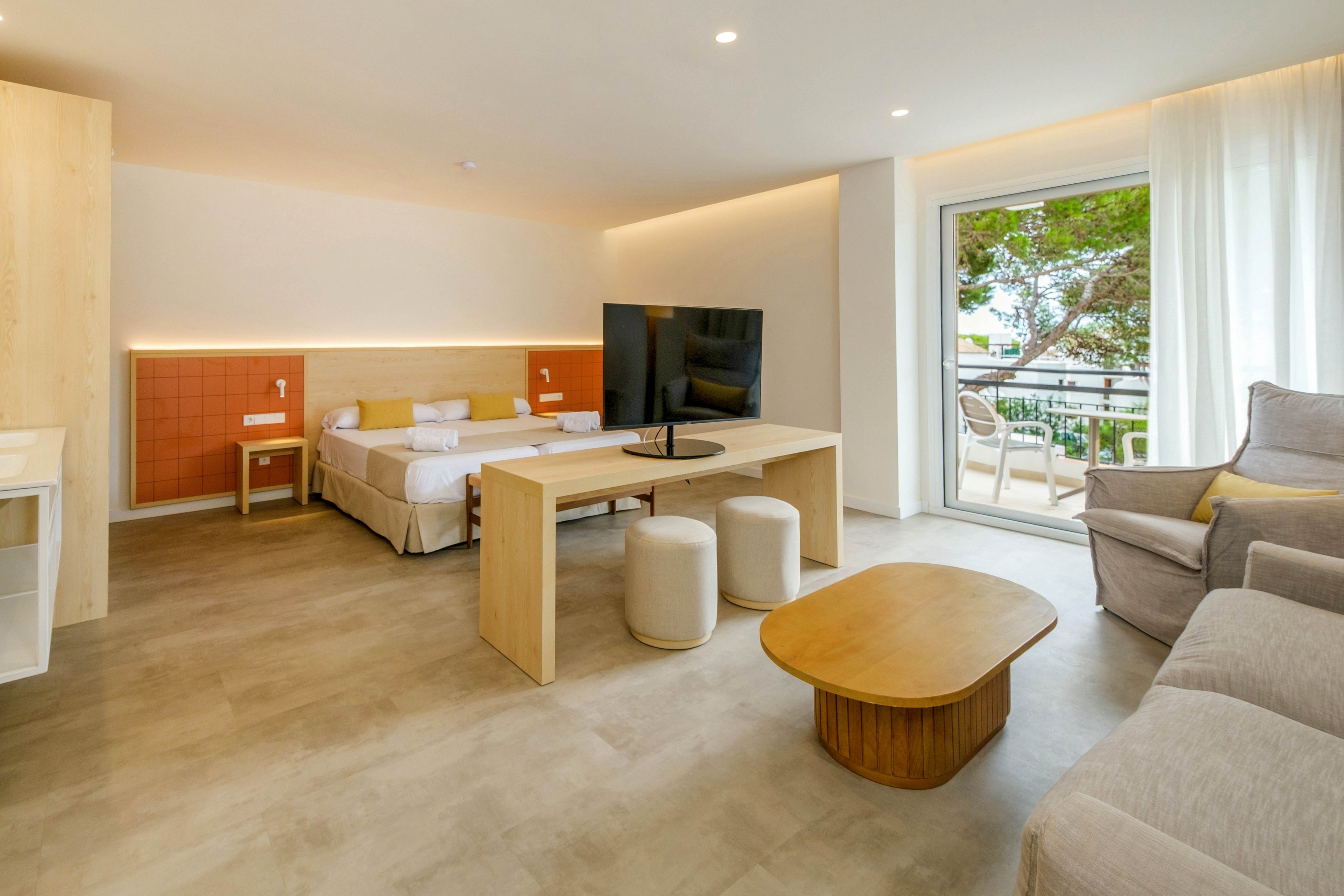 A spacious modern hotel suite features a double bed, a large flat-screen television, and minimalist wooden furniture. The room includes a desk-like divider with stools, a coffee table, and a comfortable sofa area. Floor-to-ceiling curtains frame a glass door that opens onto a balcony with outdoor seating and a view of trees. Neutral tones and soft lighting create a bright, relaxing atmosphere suitable for leisure or business stays.