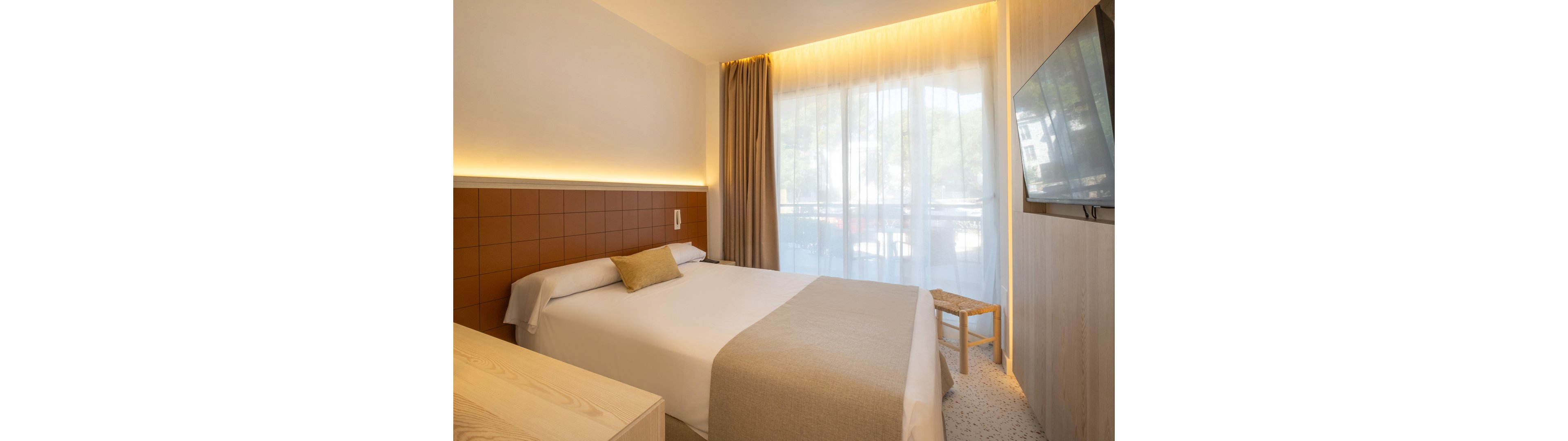 A contemporary hotel bedroom features a neatly made double bed with neutral bedding and a single accent pillow. Warm indirect lighting runs along the headboard wall, highlighting the clean, minimalist design. Floor-to-ceiling sheer curtains reveal a balcony with trees visible outside, bringing natural light into the room. A flat-screen television is mounted on the wall opposite the bed, and a simple wooden stool and light wood furniture complete the calm, modern aesthetic.