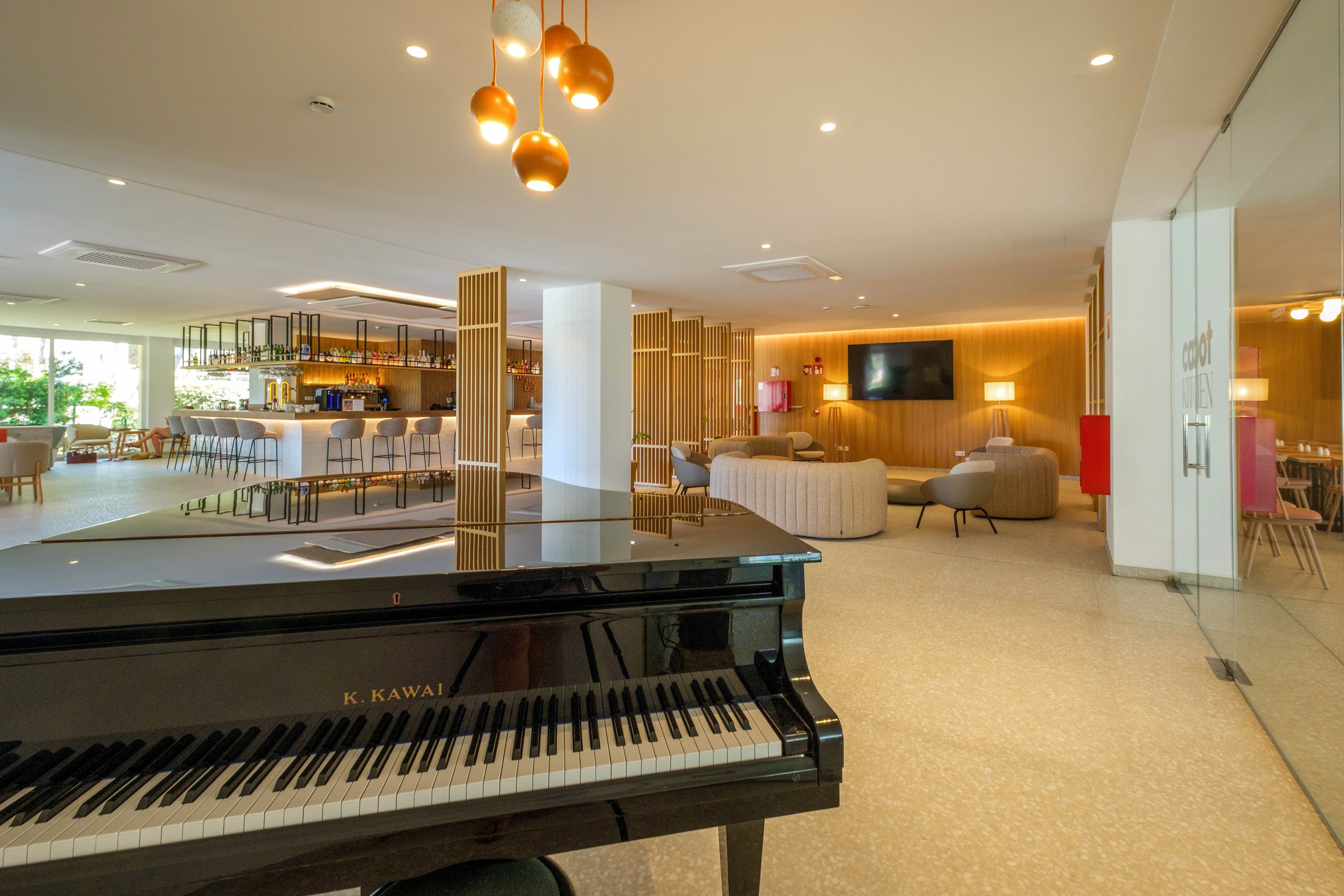 A black grand piano dominates the foreground of a contemporary lounge space. In the background, a stylish bar area with high stools and warm wooden accents creates a welcoming atmosphere. Soft seating, a large wall-mounted television, and decorative lighting add to the modern, upscale feel. Glass doors and bright ambient lighting contribute to the open, airy design.